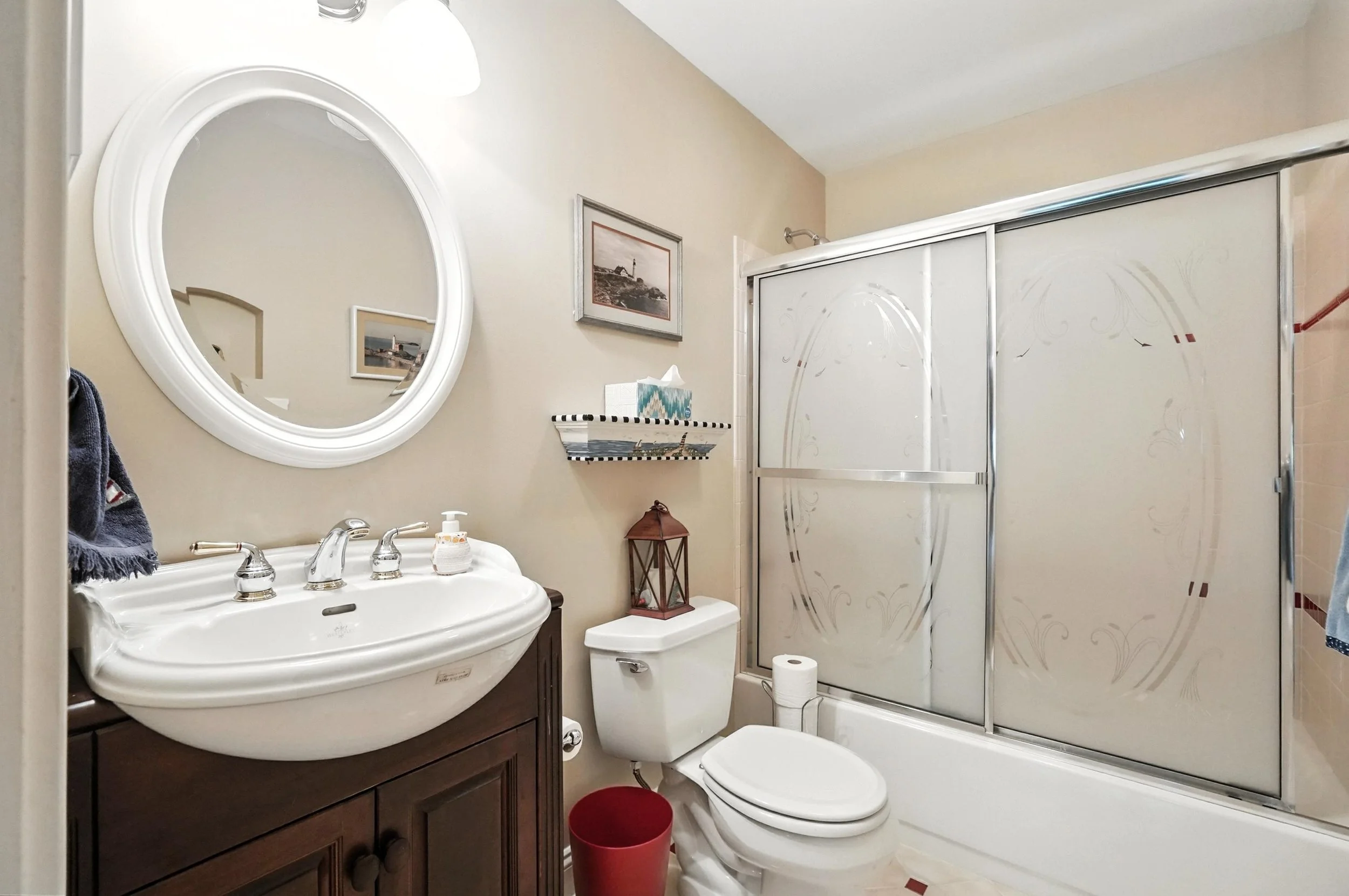 Bathroom with white sink and faucet, oval mirror, beige walls, toilet with a lantern and tissue paper on top, shower with frosted glass doors, and decorative wall art.
