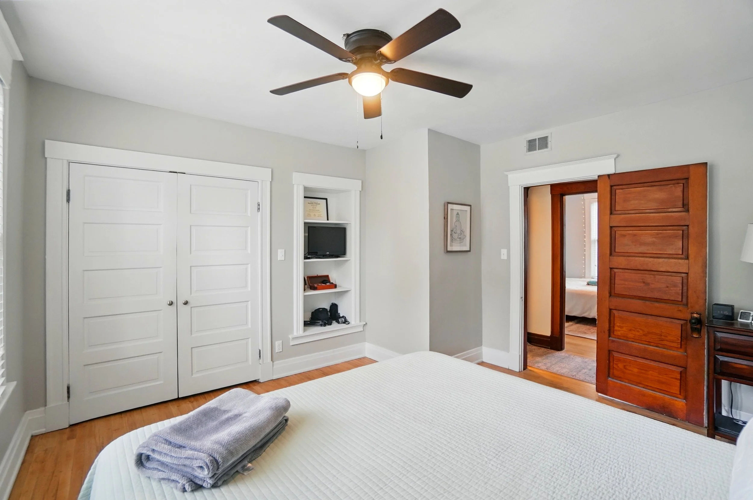A bedroom with a ceiling fan, a closed double closet, a built-in shelf with a small TV, a wooden door leading to another room, and a bed with folded towels on top.