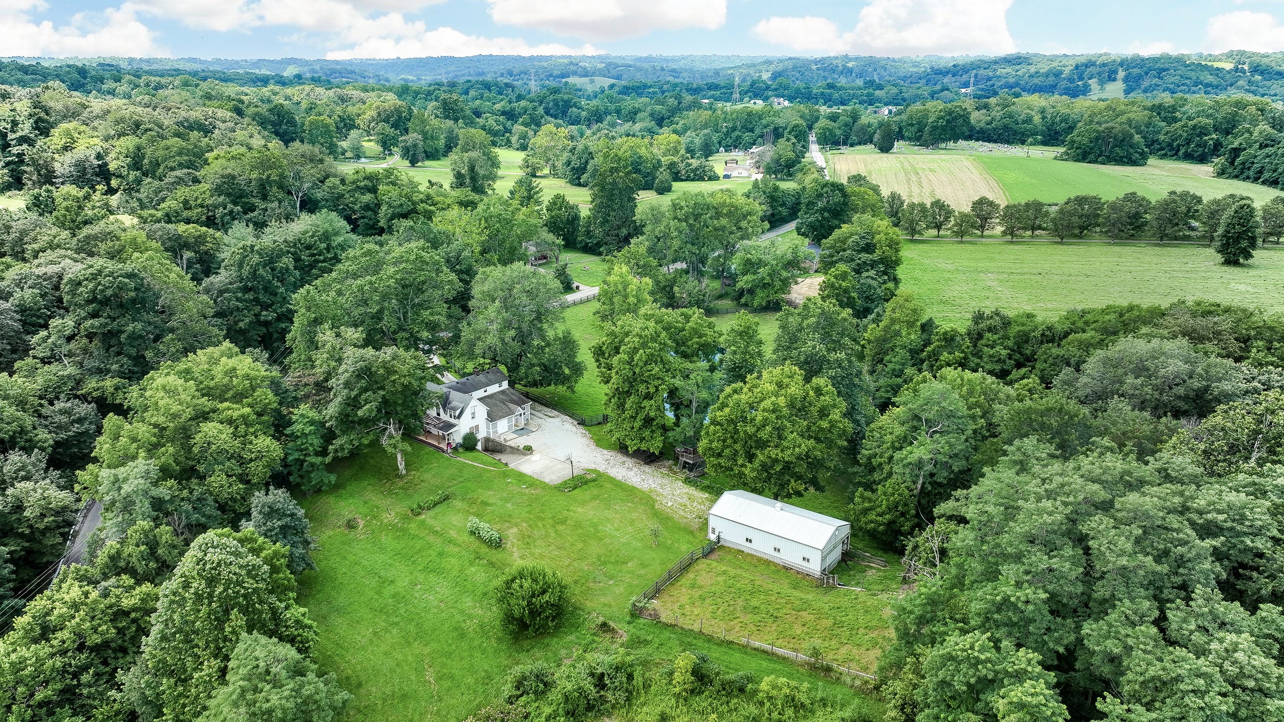 Aerial view of a countryside landscape with green trees, farmland, and a white house with a driveway, surrounded by lush green grassy areas and wooded regions.