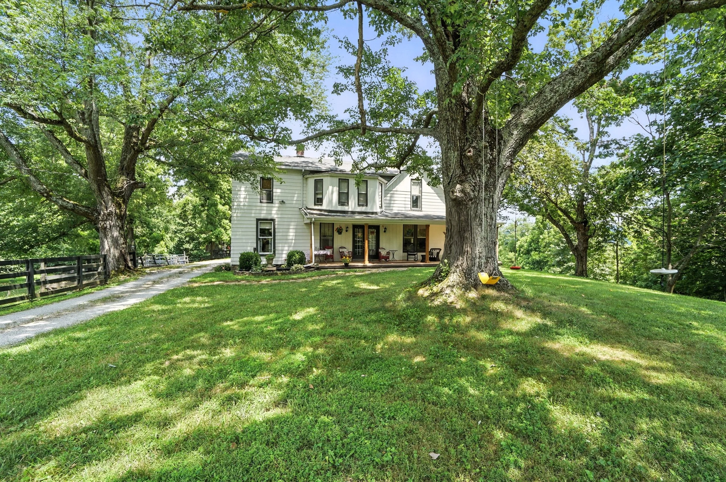 A white two-story house with a porch, surrounded by large green trees and a grassy lawn, with a gravel pathway on the left side.