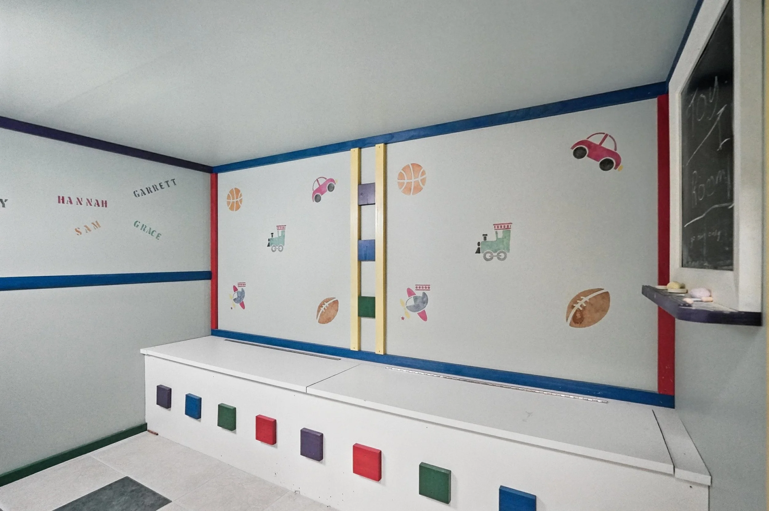 Indoor kids' play area decorated with colorful sports-themed wall stickers, including basketballs, footballs, toy cars, and trains, with a small chalkboard on the right side.