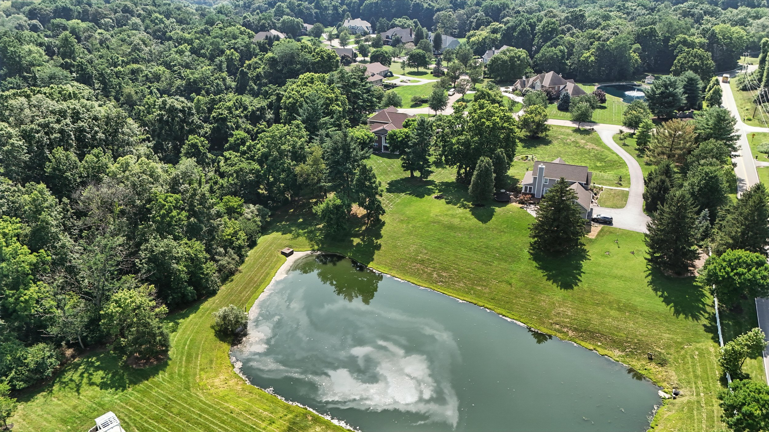 Aerial view of a neighborhood with houses and lush green trees surrounding a small pond with a boat docked nearby.