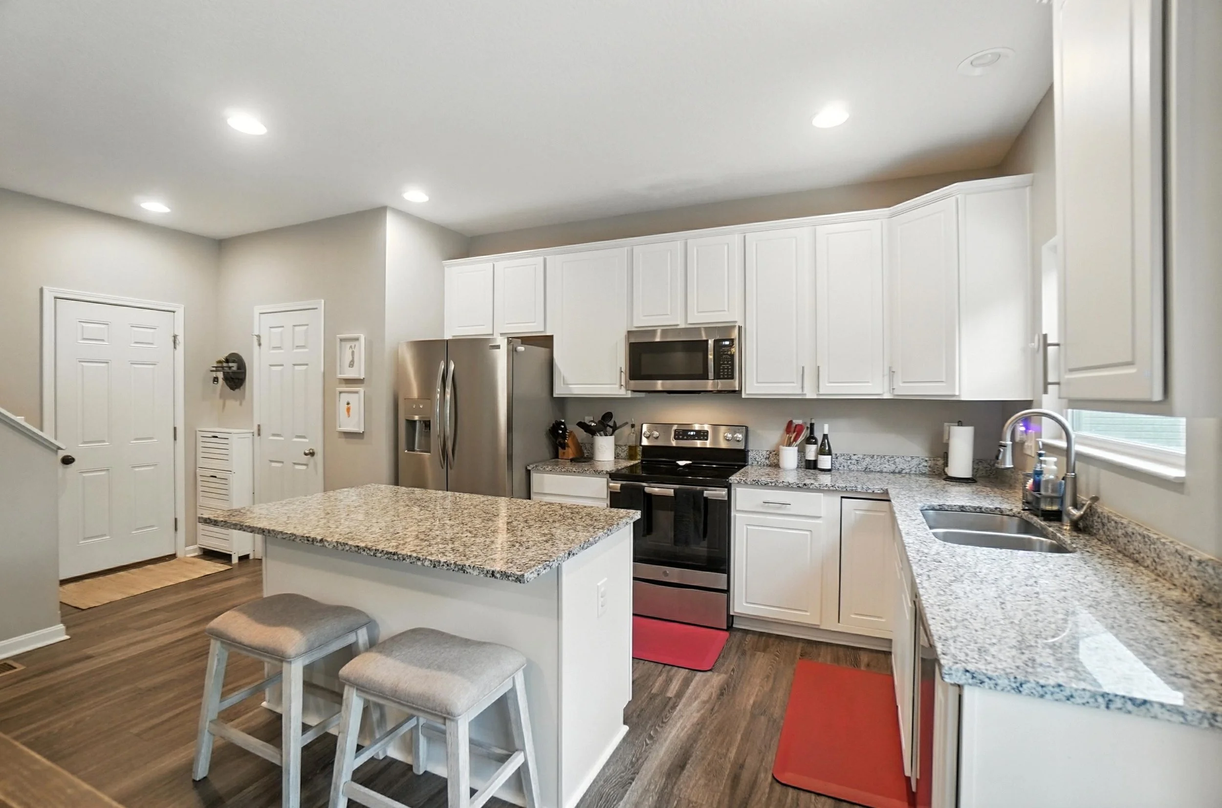 Modern kitchen with white cabinetry, granite countertops, stainless steel appliances, a kitchen island with two bar stools, hardwood flooring, a window above the sink, and various small kitchen items.