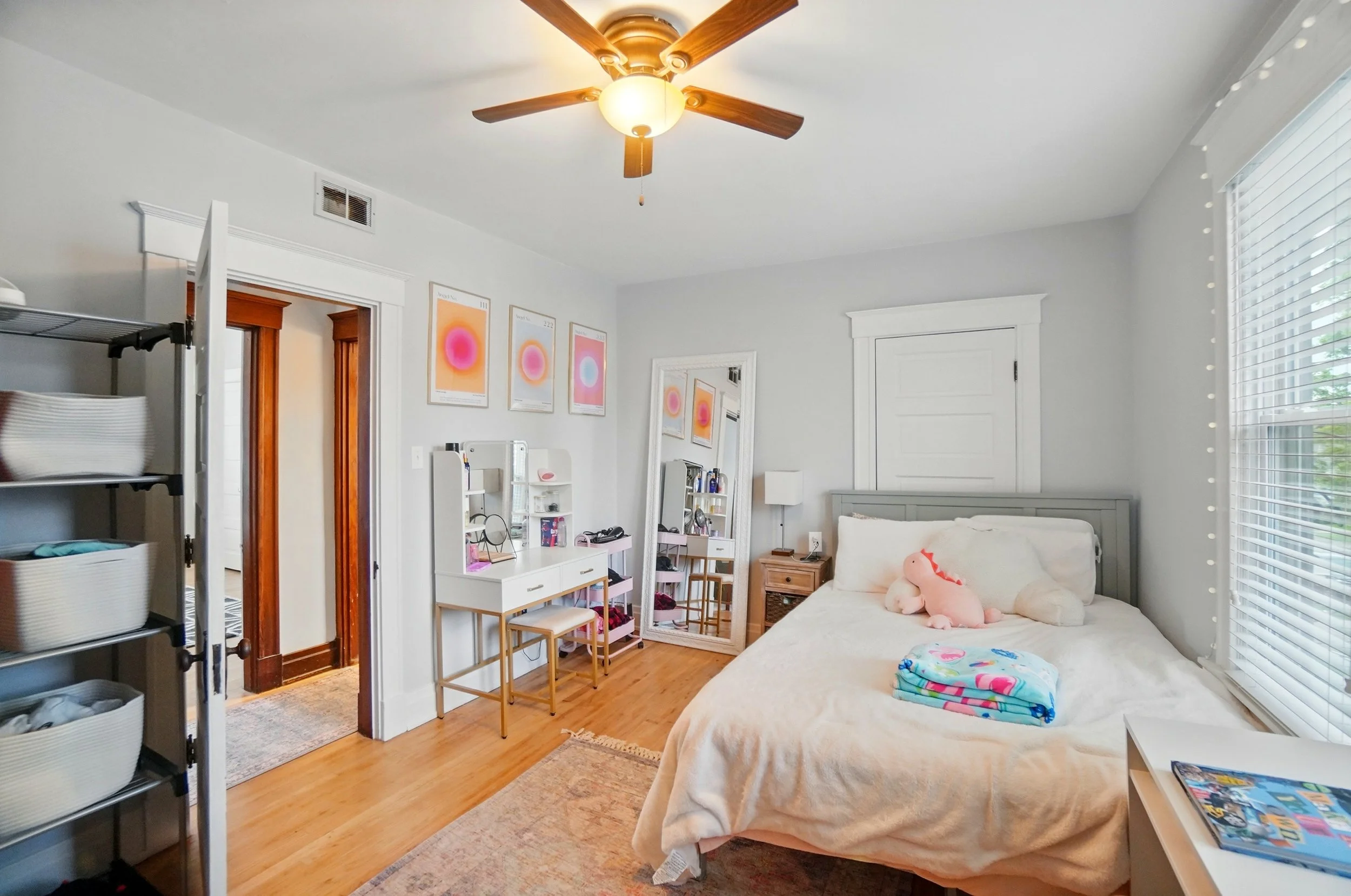 A bedroom with a bed against the wall, a window with blinds, a ceiling fan, a mirror, and various decorations.