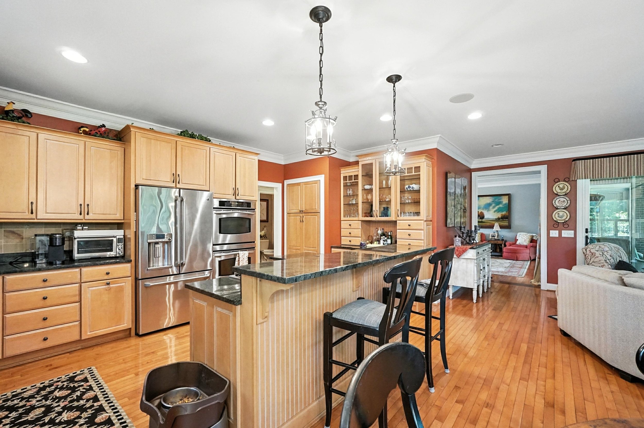 Open-concept kitchen with wooden cabinets, stainless steel appliances, a breakfast bar with barstools, and adjacent living room with sofas and artwork on the wall.