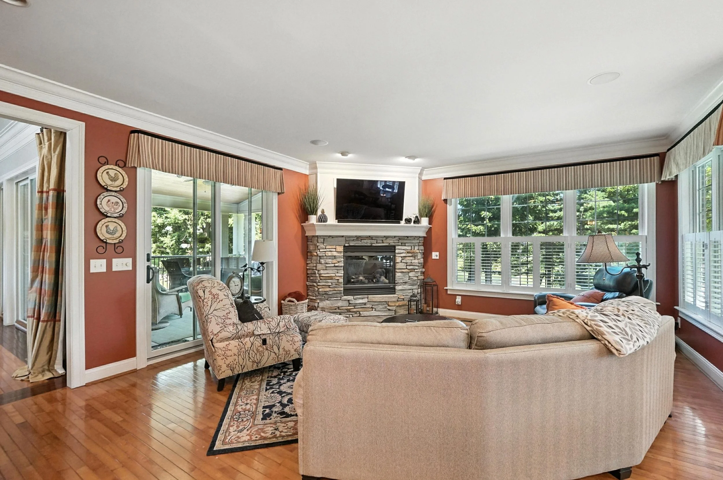 Living room with beige sectional sofa, floral armchair, stone fireplace, TV above fireplace, large windows with blinds, hardwood floors, and wall art.