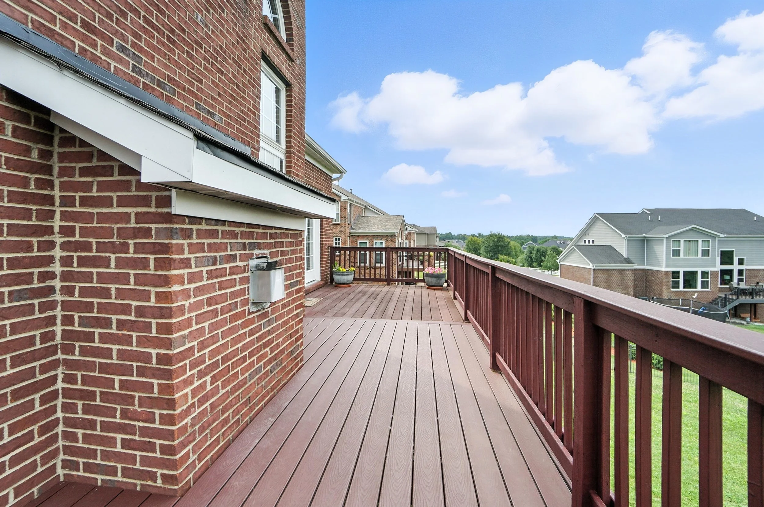 View of a spacious wooden deck with a red railing, attached to a brick house, overlooking neighboring houses and a green landscape under a partly cloudy sky.