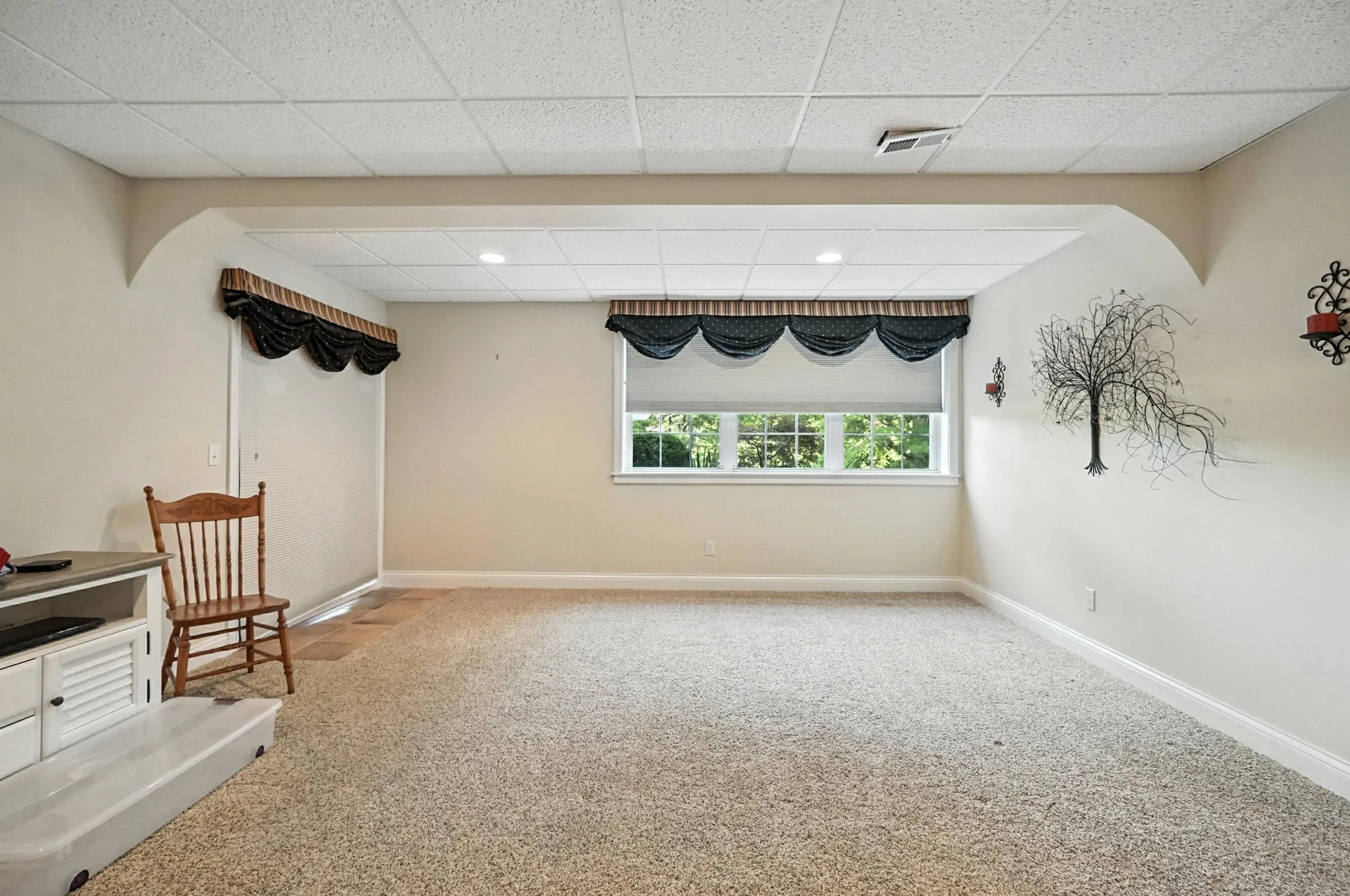 Empty living room with beige walls, window with black and beige valance, carpeted floor, wall decoration of a black wire tree, and small wall sconces with red candles.