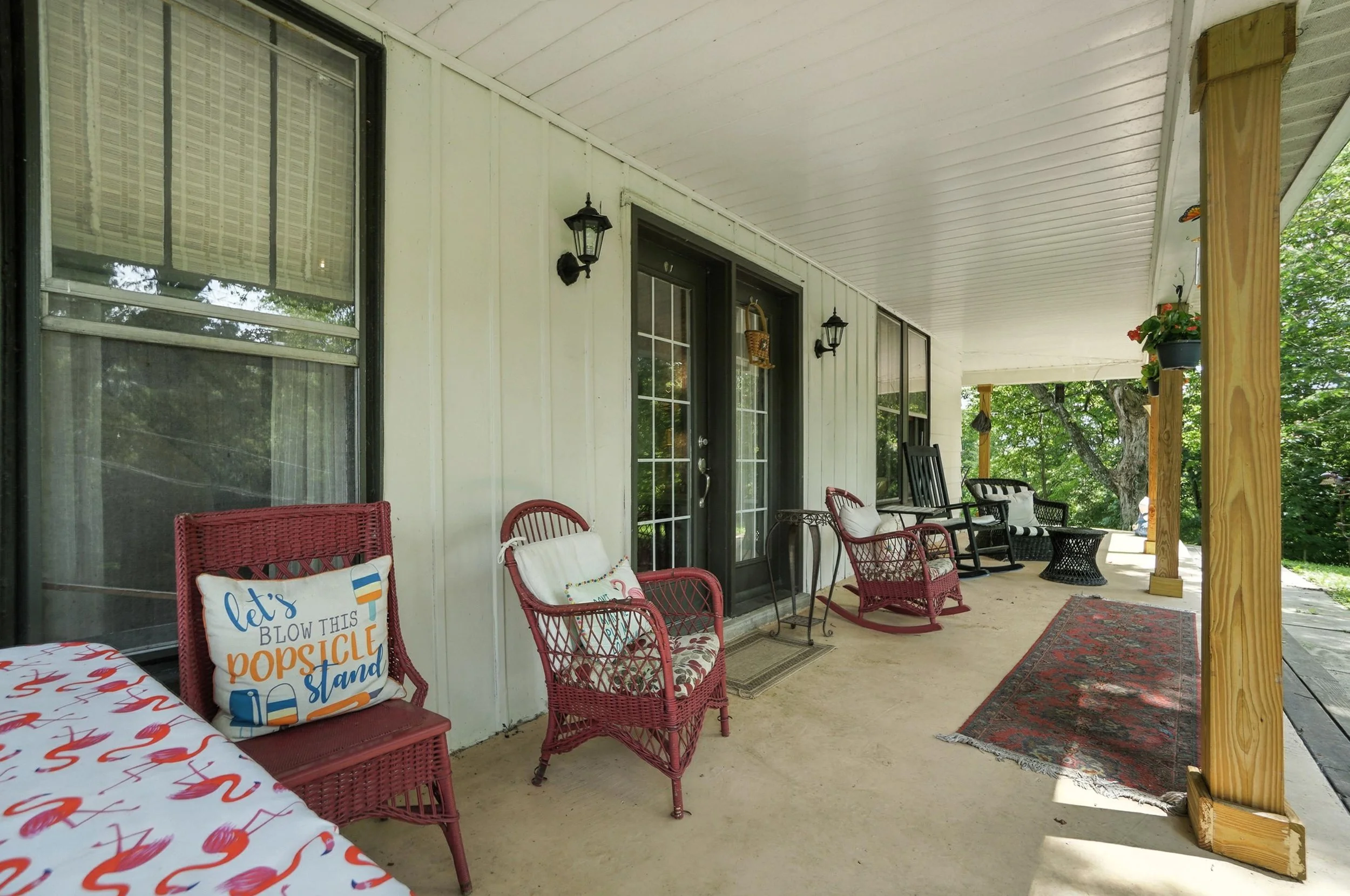 Covered front porch with wicker chairs, outdoor cushions, a rug, potted plants, and trees in the background.