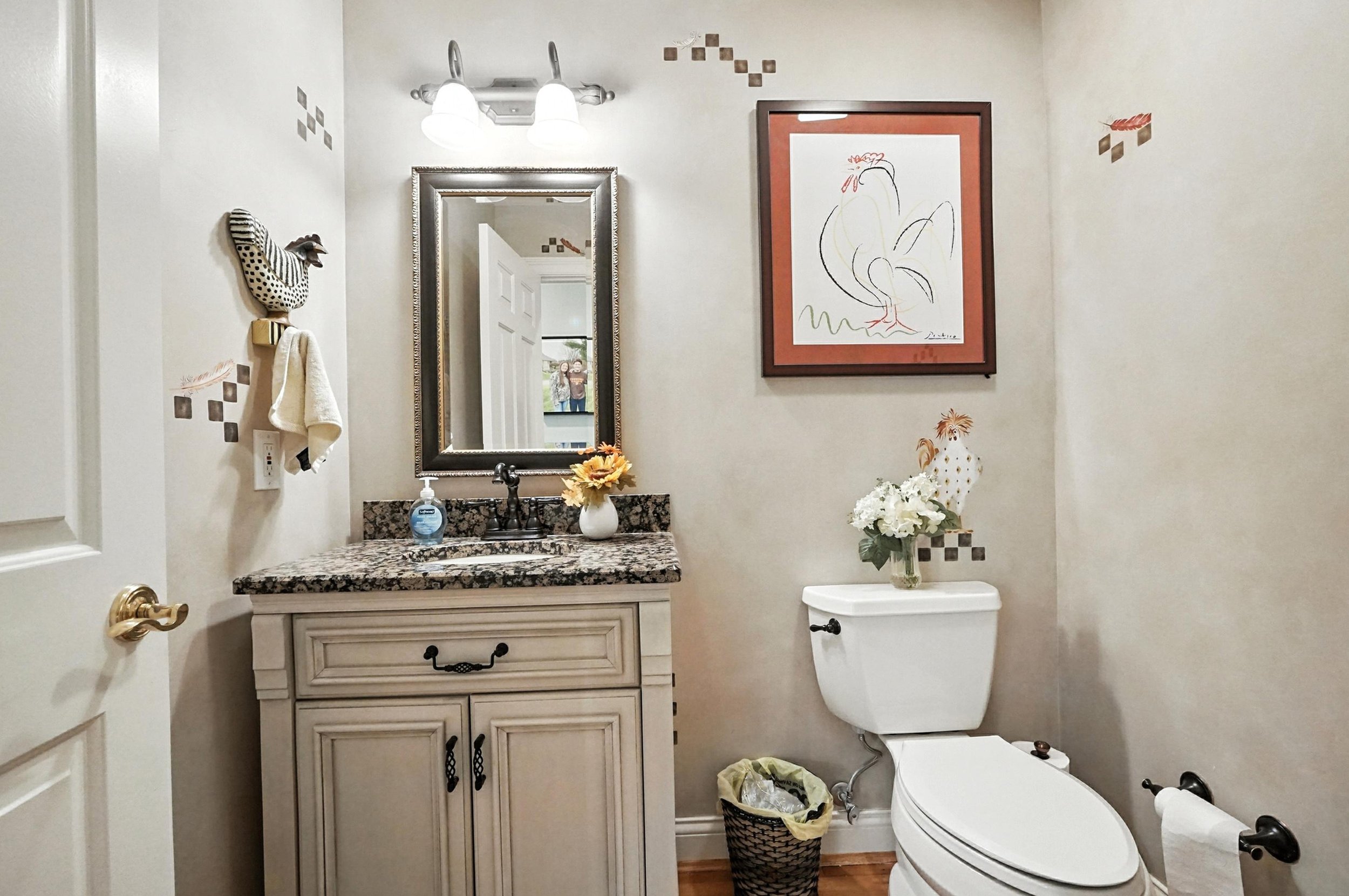 A bathroom with a vanity that has a granite countertop, a mirror, and a small vase with sunflowers. The toilet has a vase with white flowers on top. The wall has farm animal artwork and decorative tiles, with a towel and a chicken-shaped wall hook.