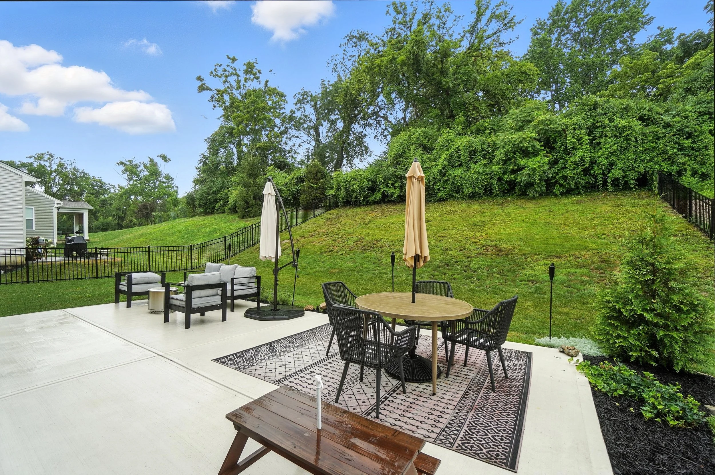 Backyard patio with patio furniture including a round table with four chairs, umbrellas, and a seating area with four cushioned chairs, surrounded by a grassy lawn and trees.