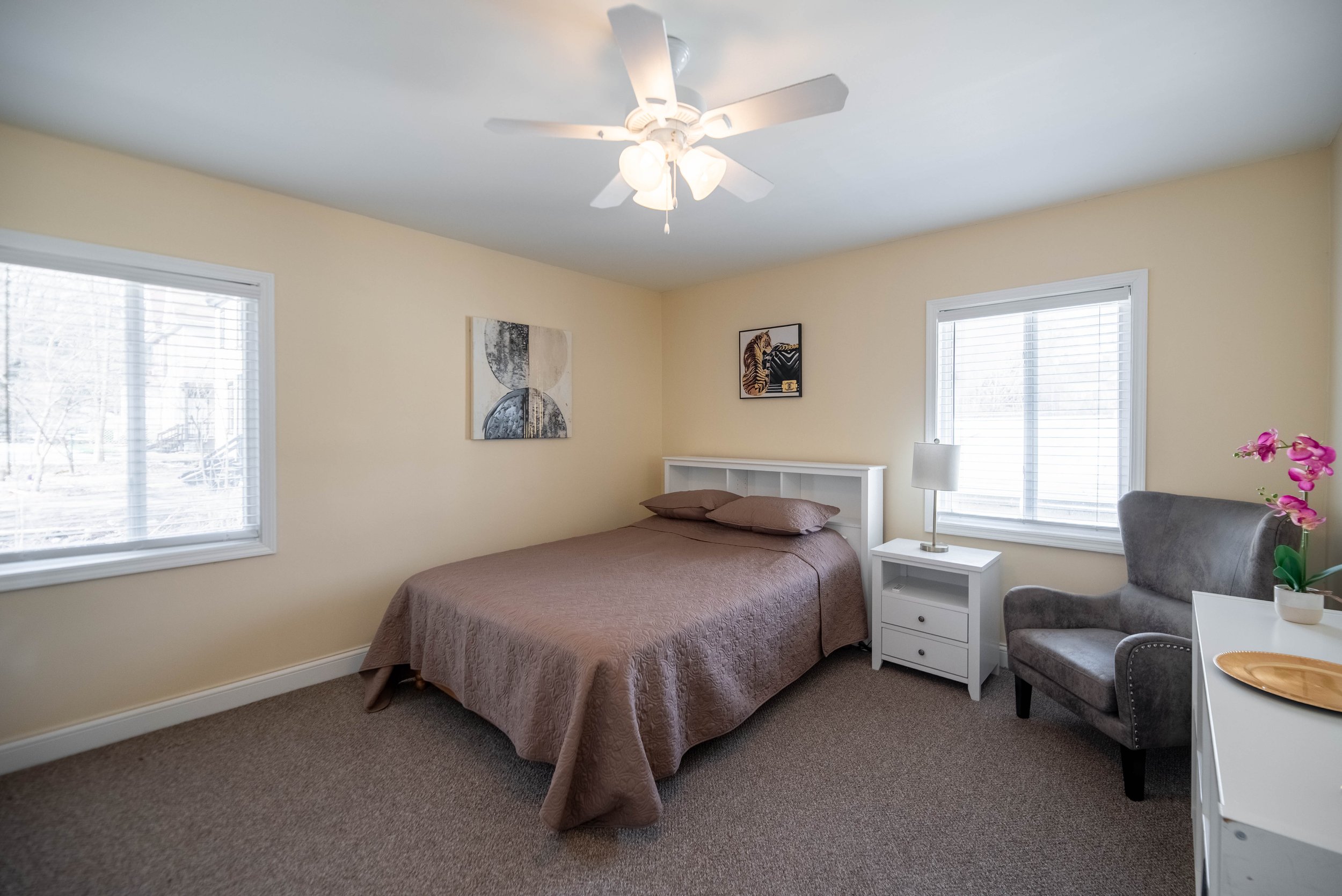 Bedroom with a bed, brown bedspread, white side table, lamp, armchair, and wall art.