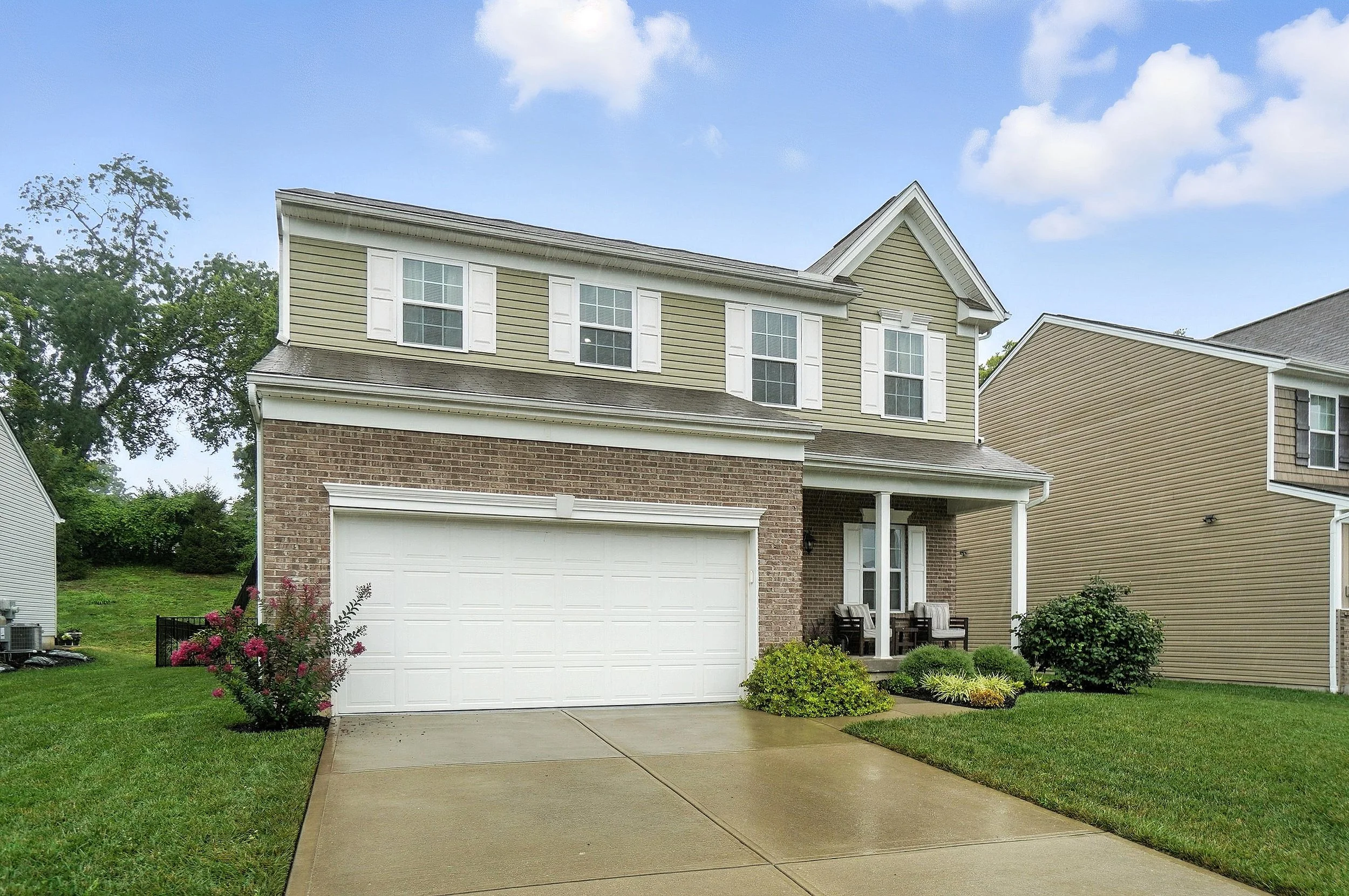 Front view of a two-story suburban house with beige and brick exterior, white shutters, a white garage door, a small front porch with two chairs, and a well-maintained lawn with bushes and flowers, under a partly cloudy sky.
