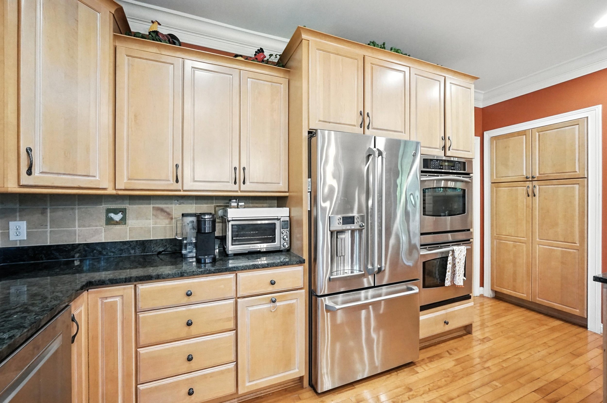 Kitchen with light wood cabinets, black countertop, stainless steel refrigerator, oven, microwave, and coffee maker, with orange painted wall and wooden floor.