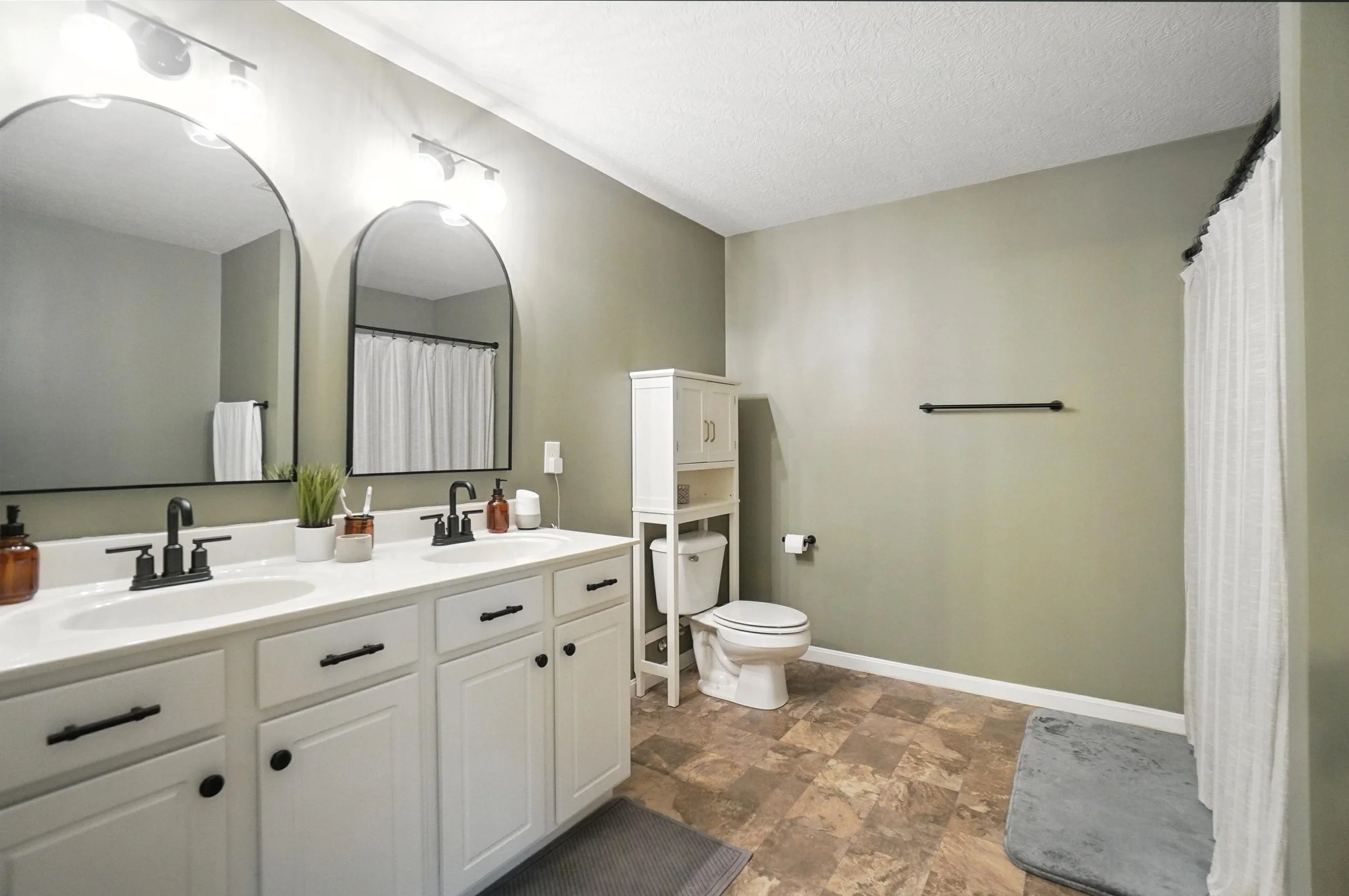 Bathroom with double vanity sinks, large mirrors, a white toilet, white cabinetry, beige walls, and a shower area with a white curtain.