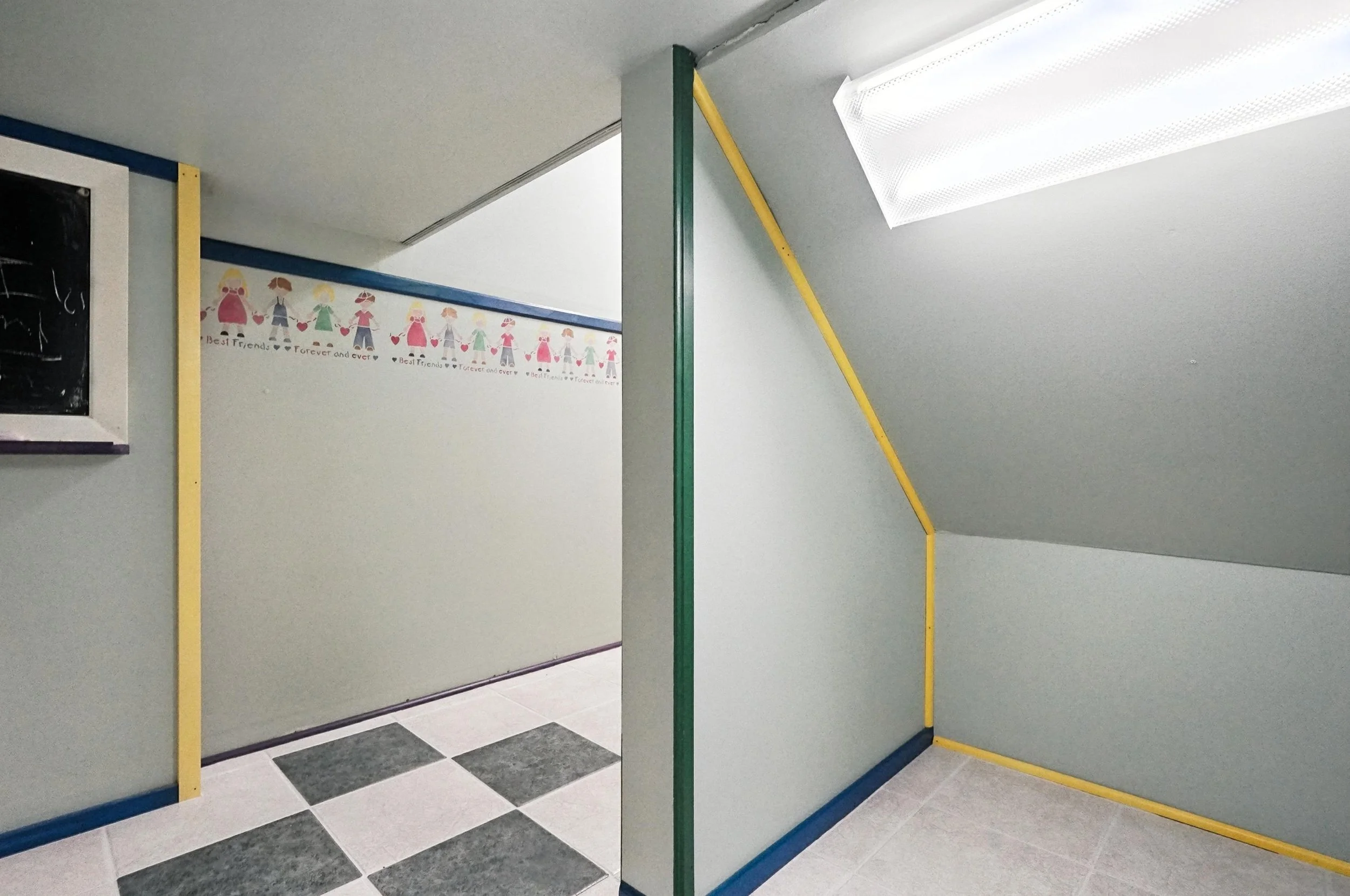 Interior of a room with a sloped ceiling, a small blackboard on the wall, a patterned border with children holding hands, and tiled flooring