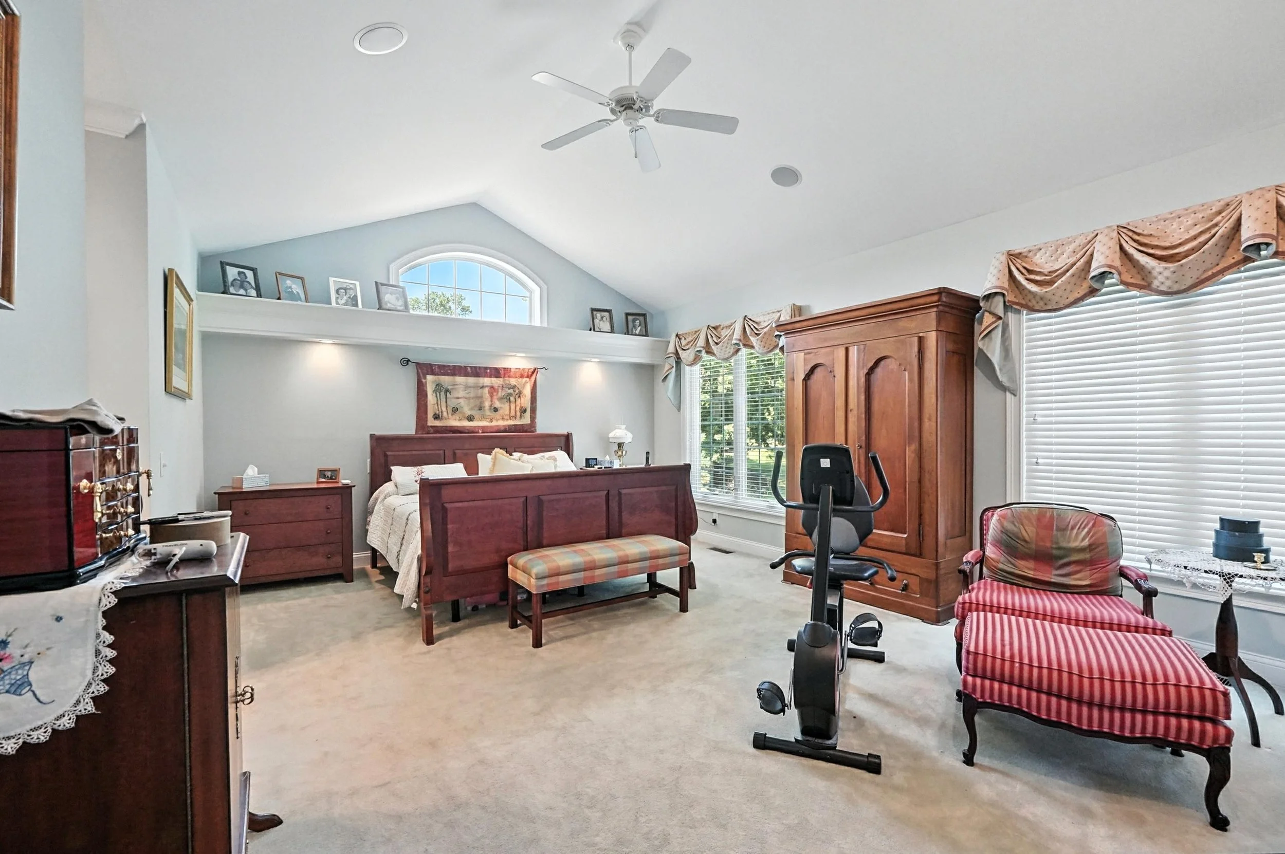 Room with a queen-sized bed, wooden furniture, exercise bike, red-striped chaise lounge, and large windows with blinds and curtains, in a house bedroom.