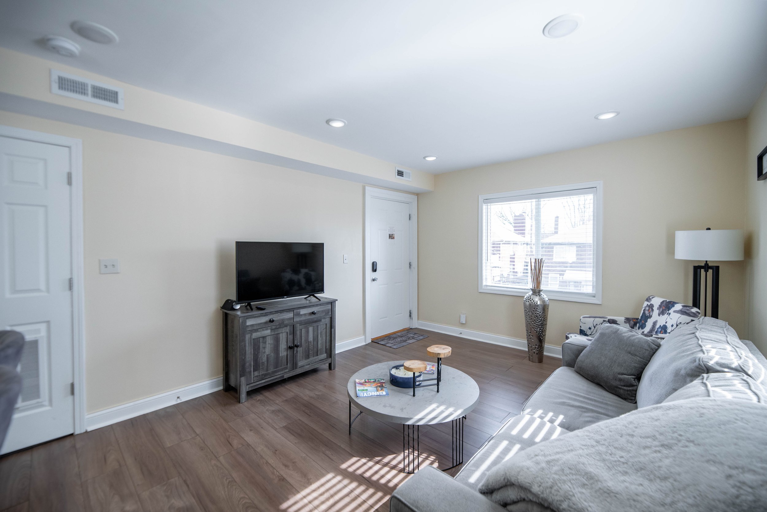 Modern living room with gray couch, TV on wooden stand, round coffee table with decor, window with blinds, and floor lamp.