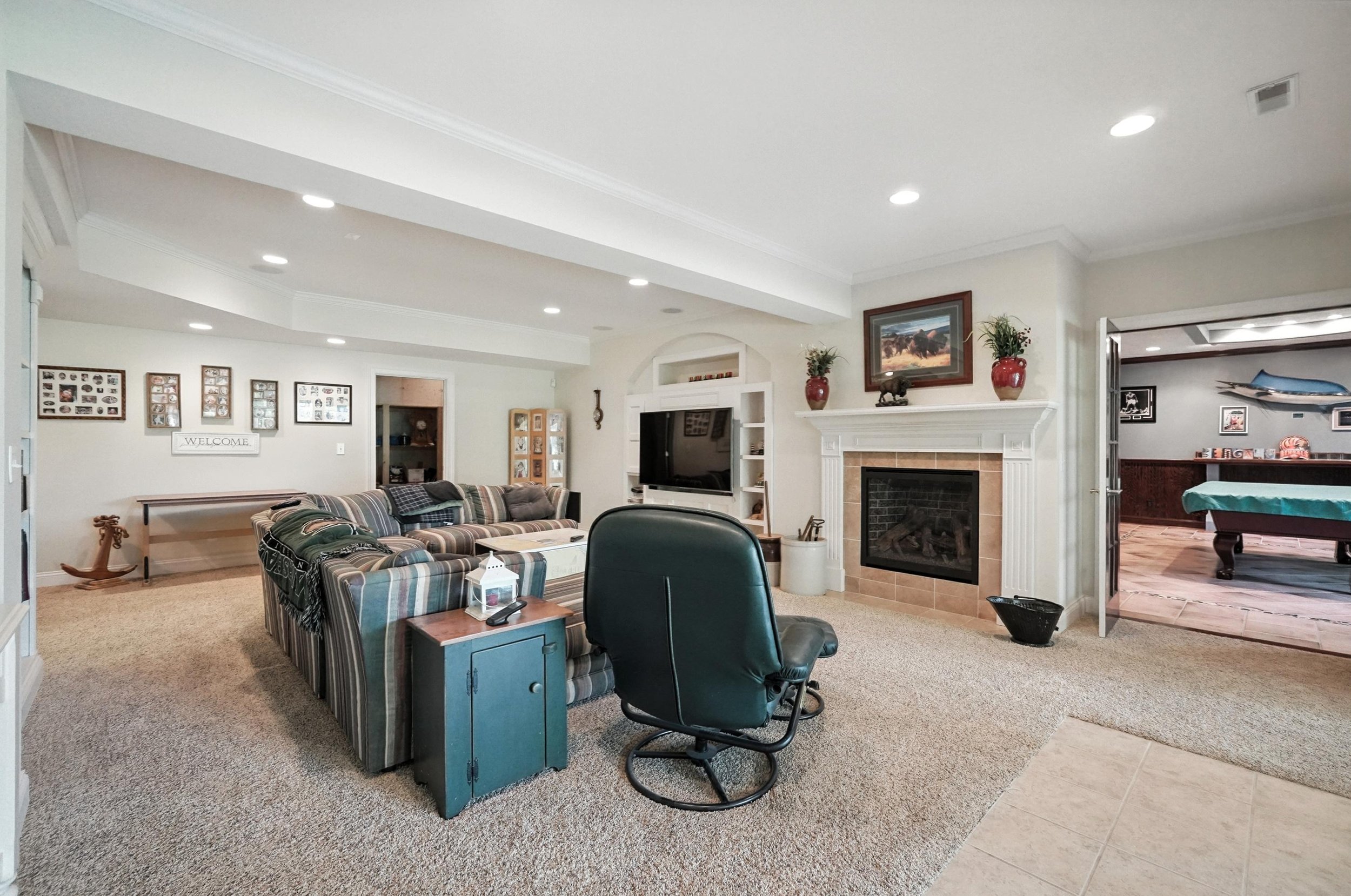 Living room with a striped sofa, a black office chair, a fireplace with a painting above it, and a flat-screen TV. There's a pool table in the adjacent room visible through an open doorway.