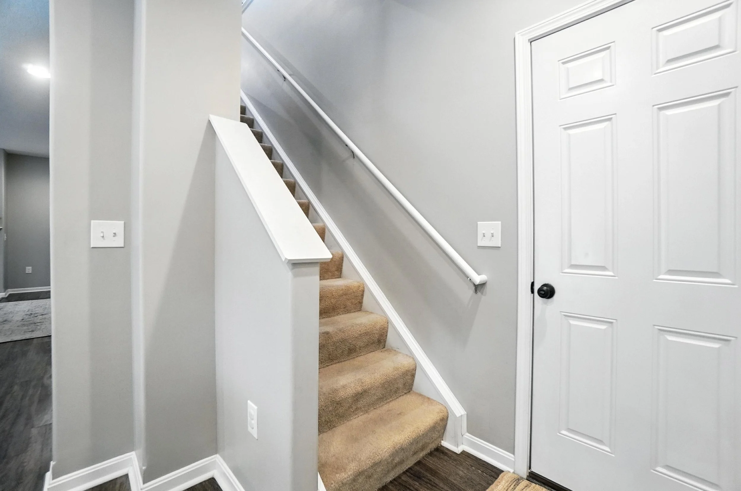 Interior view of a staircase with beige carpet leading upward, adjacent to a white door with a black doorknob, and light gray painted walls.