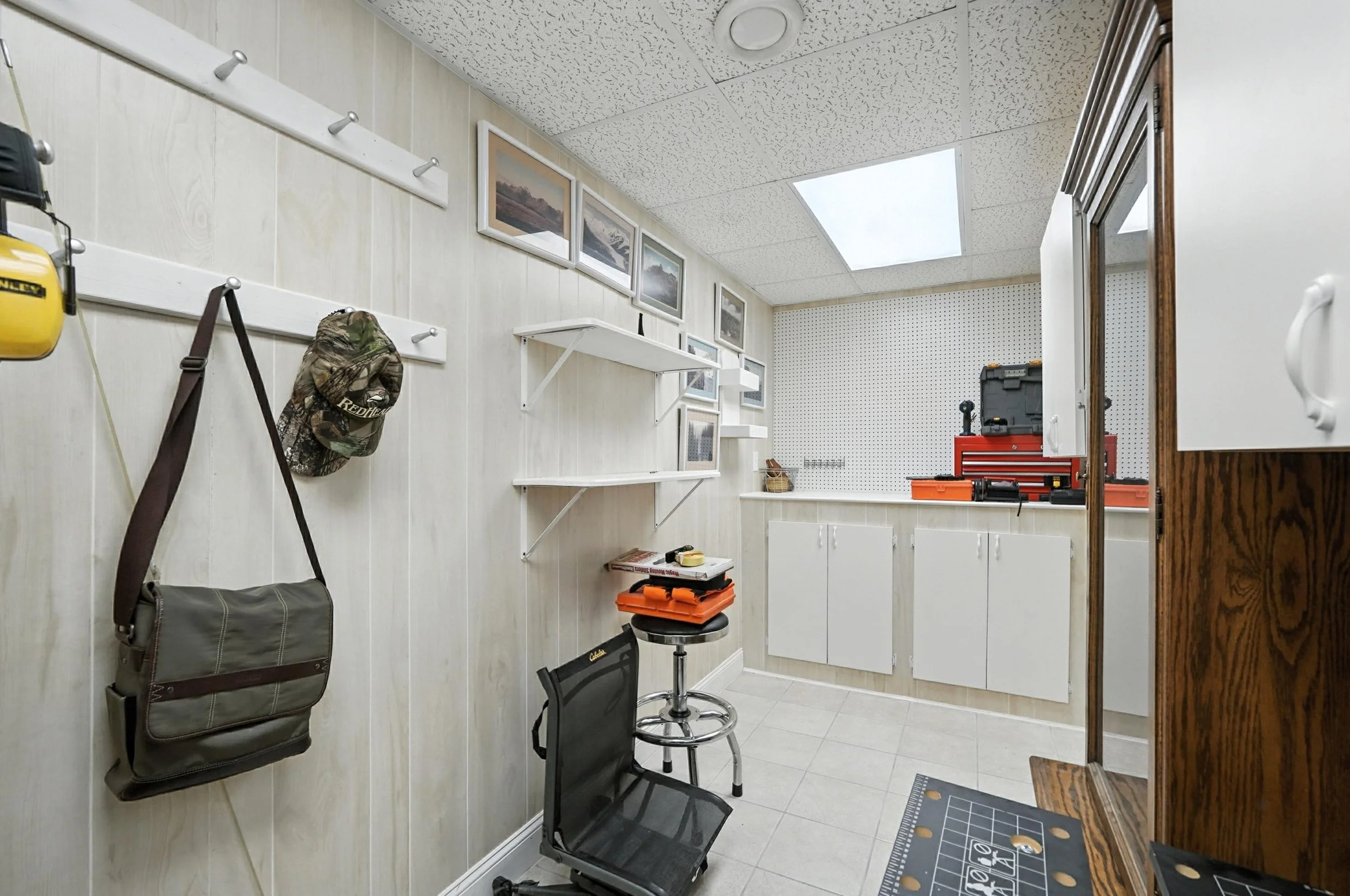 A small workshop with white paneled walls, ceiling tiles, and fluorescent lighting. There are mounted hooks with a bag and camouflage hat, framed landscape photographs, open white cabinets, a black office chair, a red toolbox, and work tools on a cou