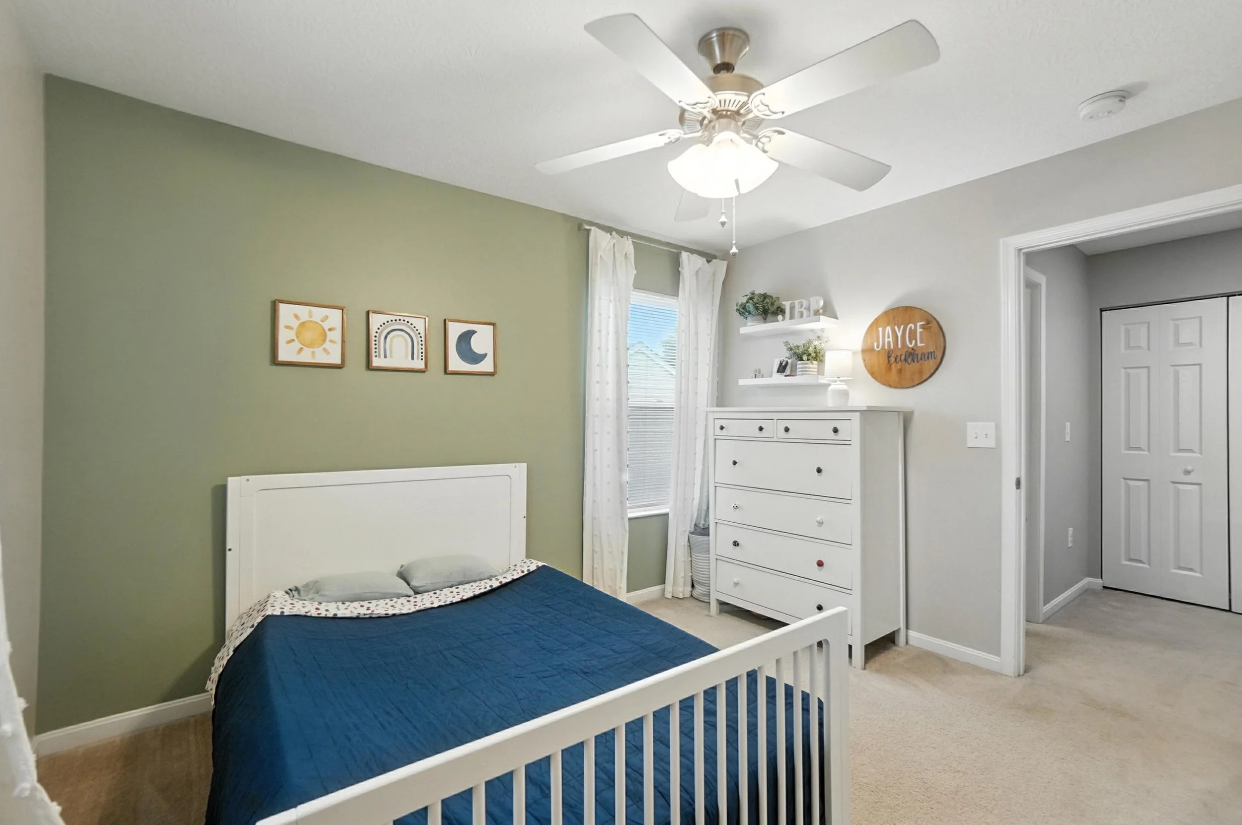 A bedroom with a green accent wall, a white bed with blue bedding, framed art of a sun, rainbow, and moon, a window with white curtains, a white dresser, decorative shelf with plants and letters, and a ceiling fan.