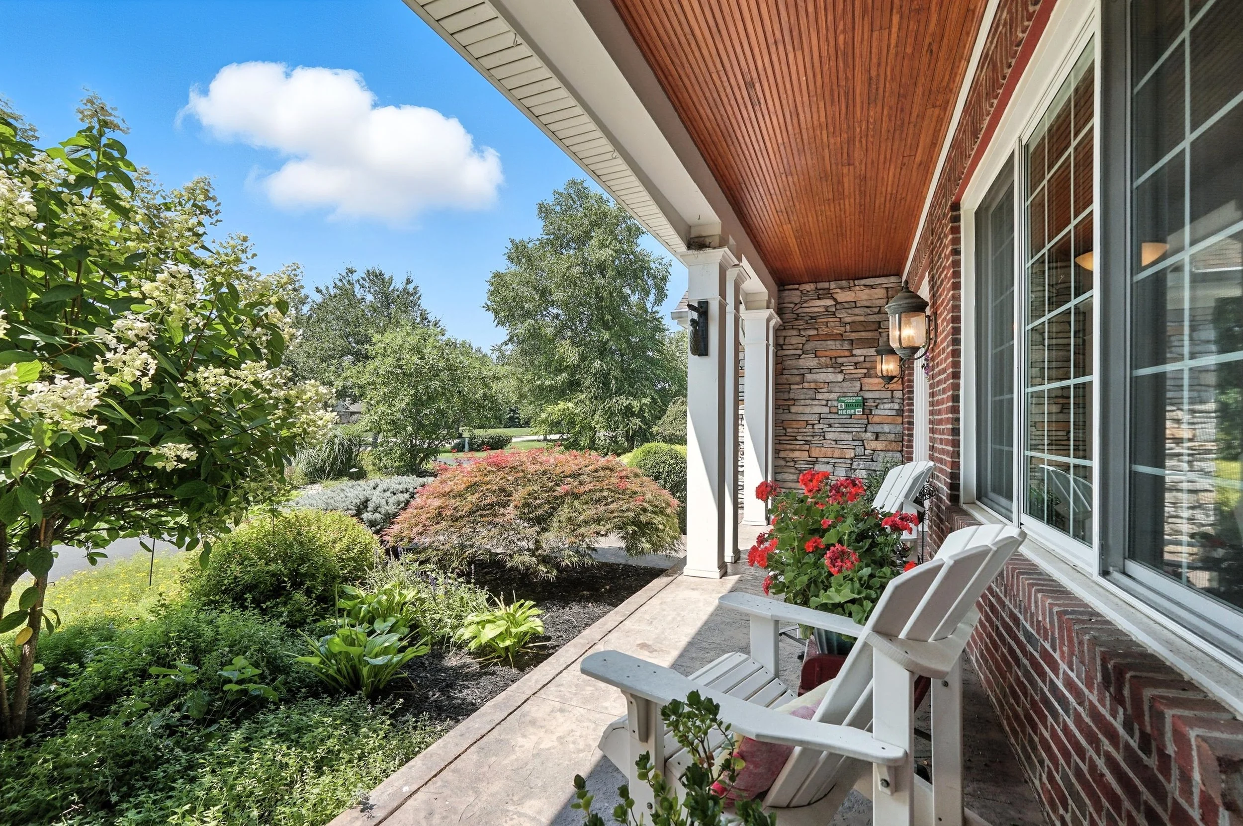 Front porch with white Adirondack chairs and red flowers, brick and stone house exterior, lush green garden with trees and bushes, blue sky with clouds.