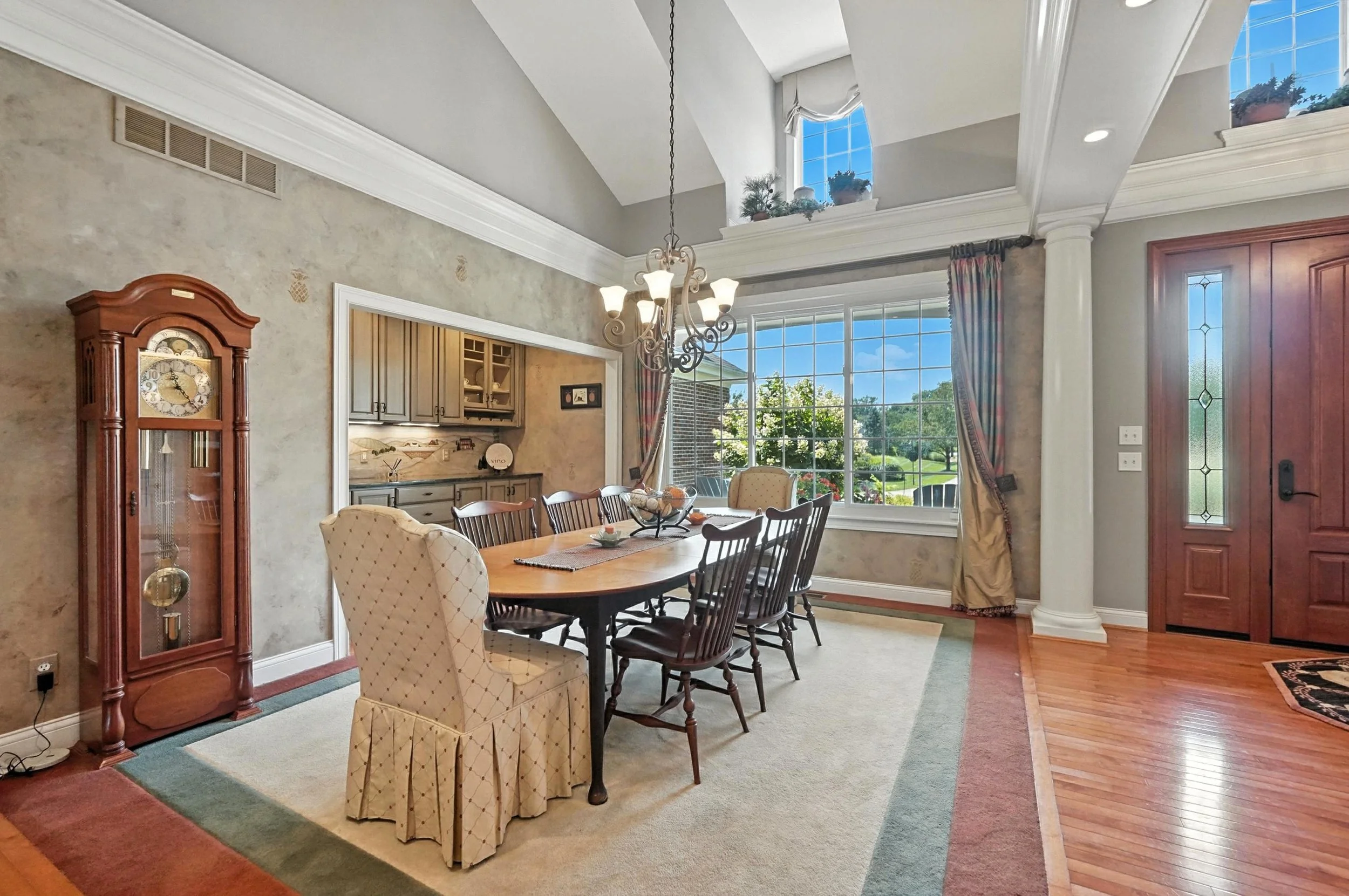 Dining room with wooden table, eight chairs, a grandfather clock, large window showing greenery outside, and a chandelier hanging above the table.