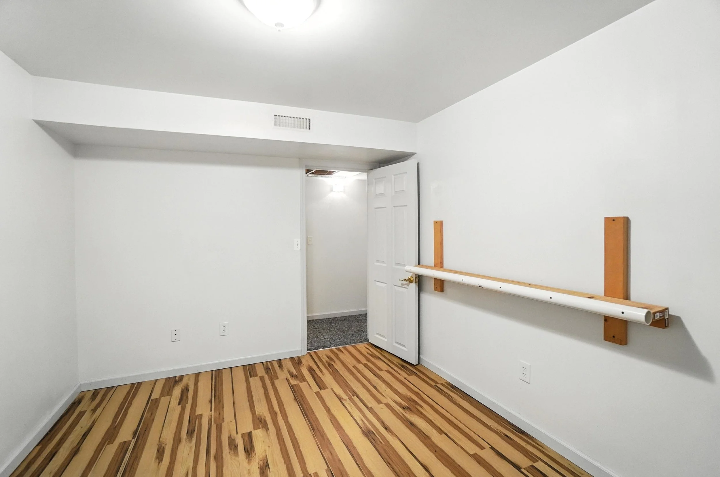 Empty room with white walls, wooden floor, and a white door. A wall-mounted handrail is attached to one wall. An open doorway leads to a carpeted area.