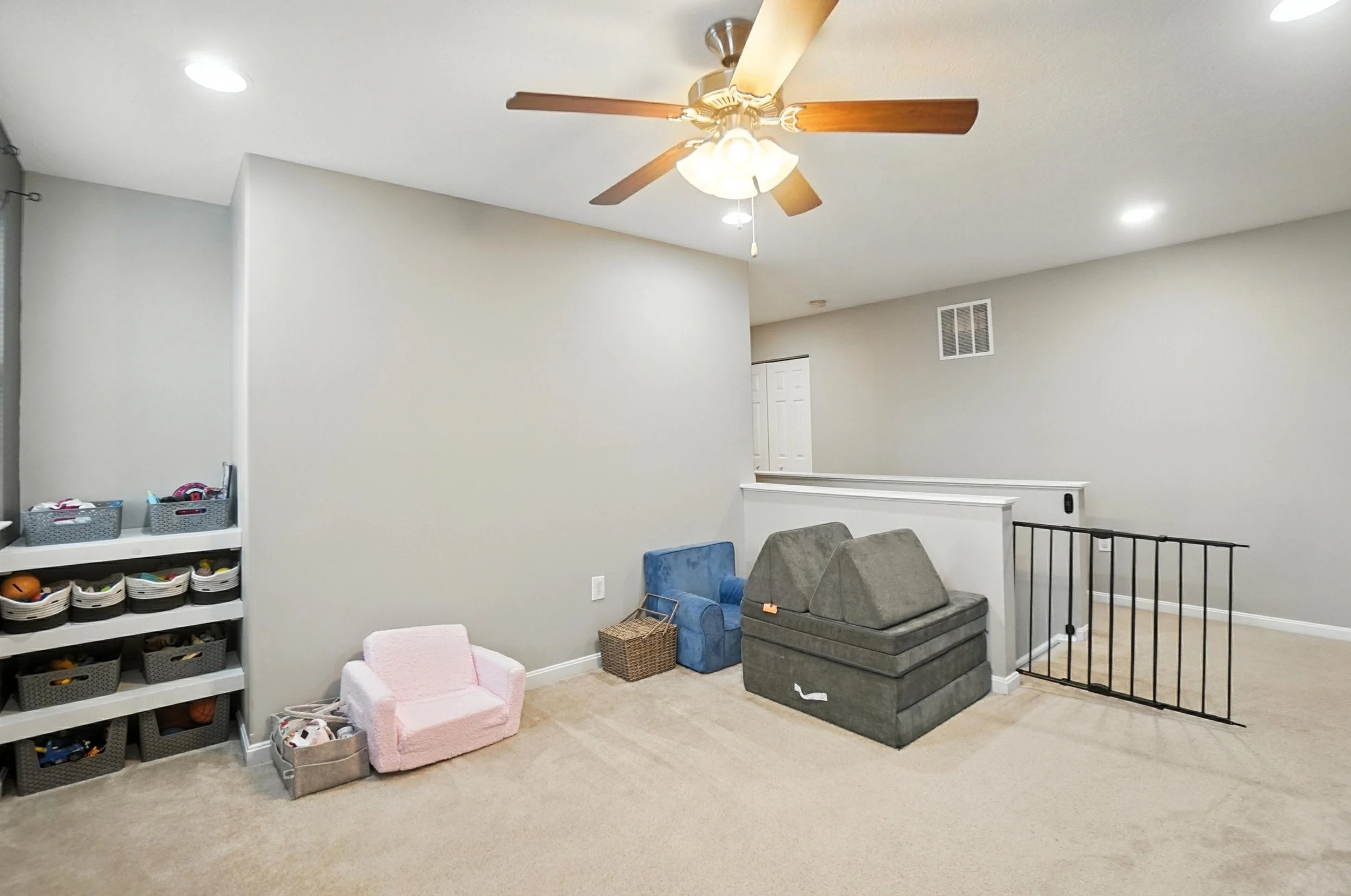 Empty living room with beige carpet, gray walls, ceiling fan with wooden blades, and various children's toys and furniture.