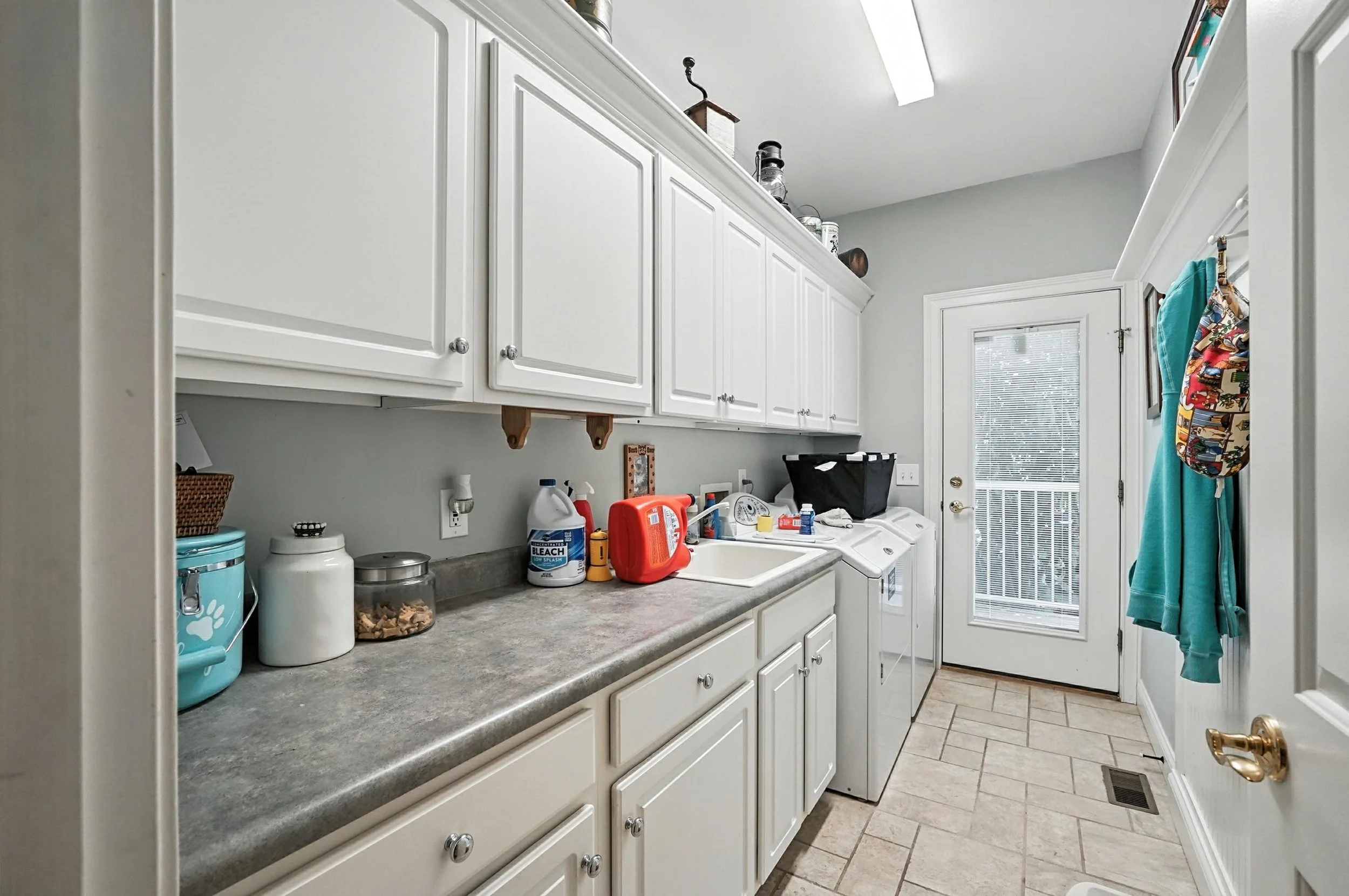 Laundry room with white cabinets, countertop with cleaning supplies, laundry machines, and a door with a window leading outside.