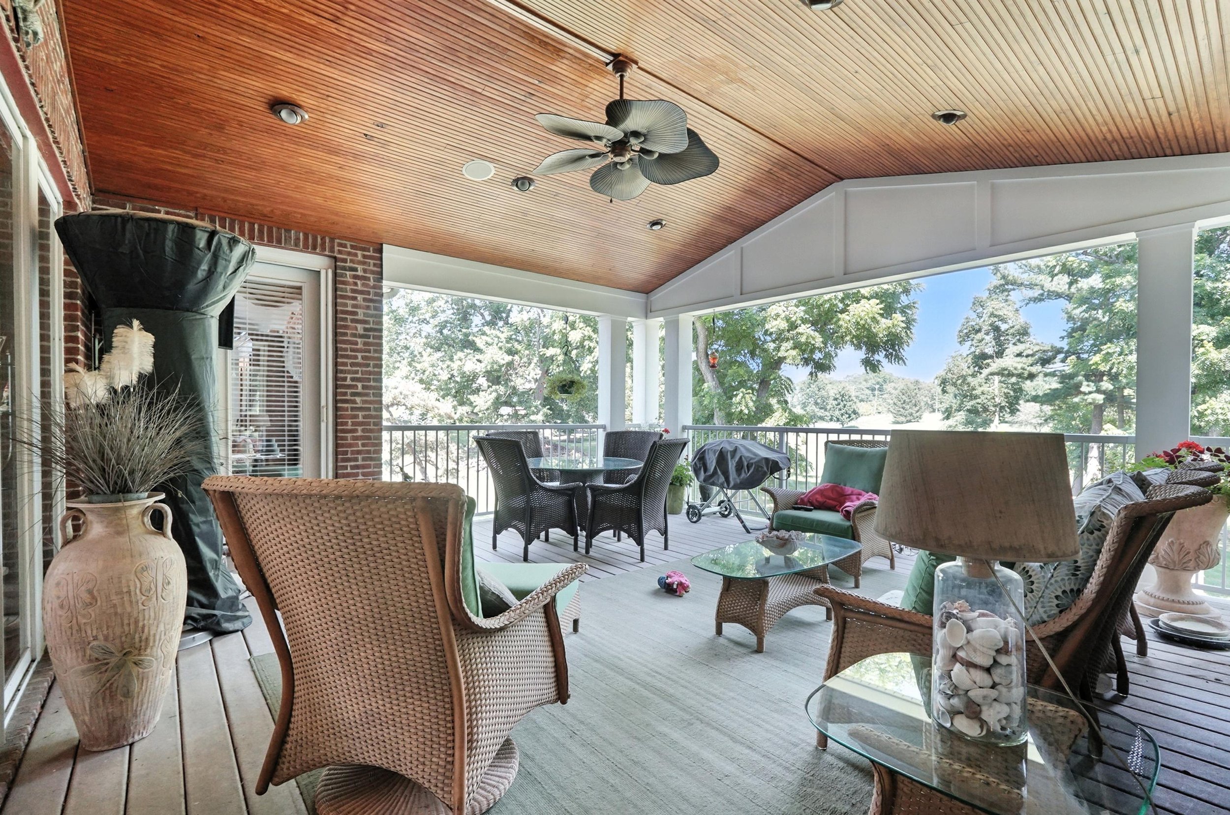 Covered screened porch with wicker and cushioned seating, a glass table, a ceiling fan, potted plants, and trees outside.