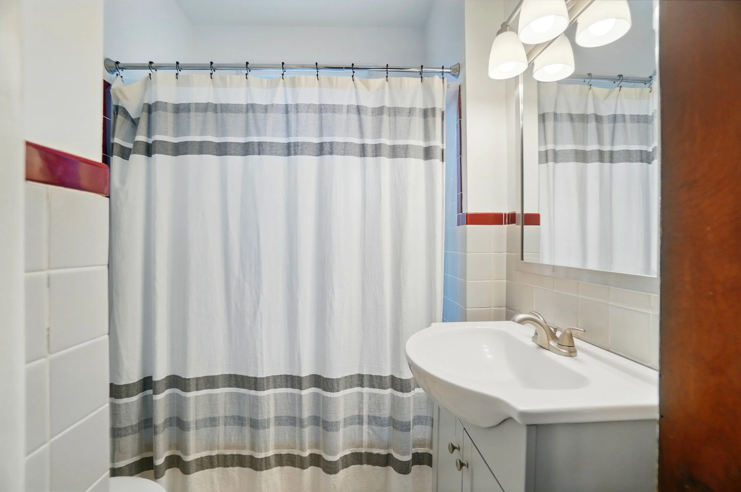 Bathroom with white sink, mirror, wall-mounted light fixture, and a shower curtain with horizontal stripes.