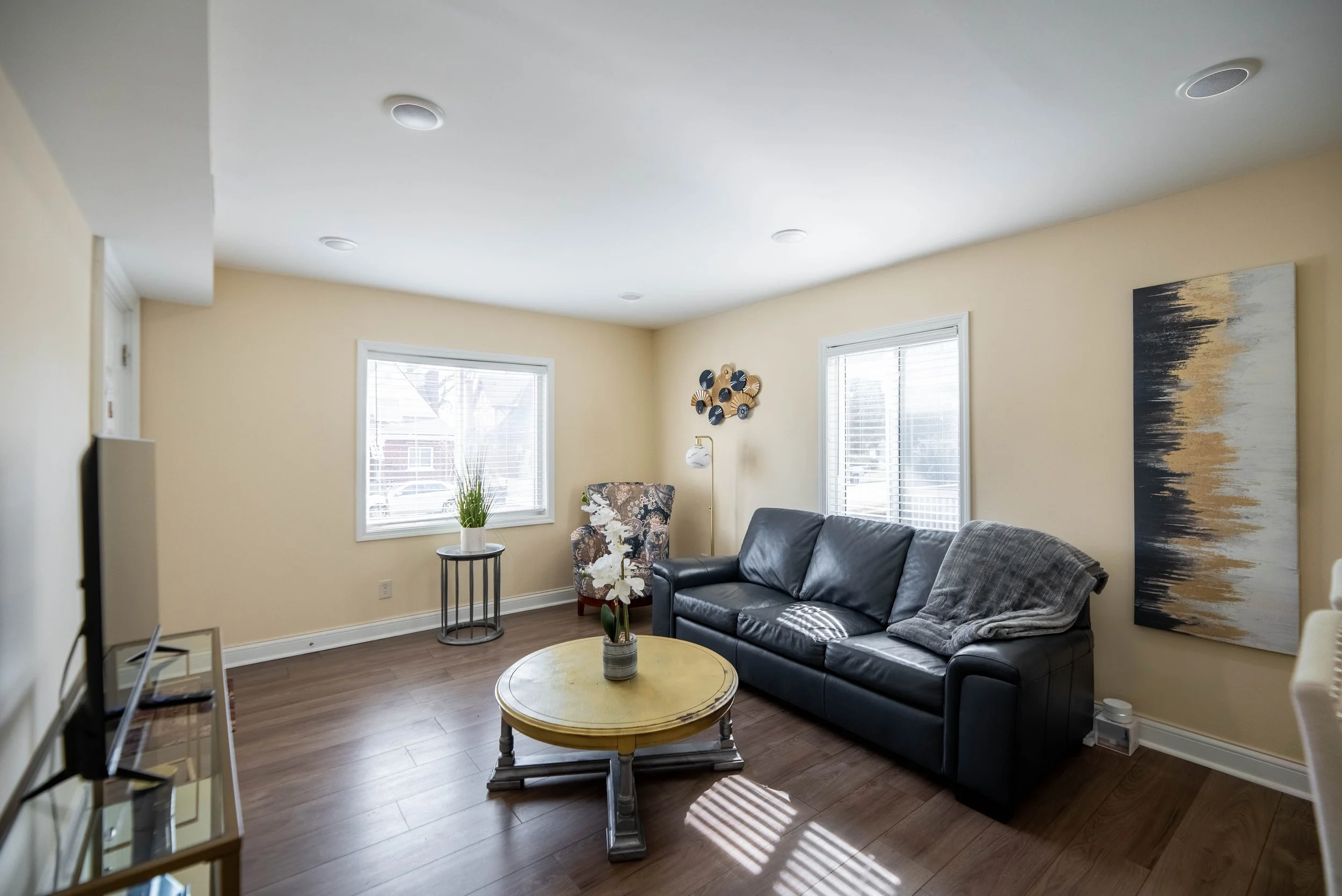 Cozy living room with beige walls, a black leather sofa, floral armchair, round wooden coffee table, abstract wall art, and large windows with blinds.