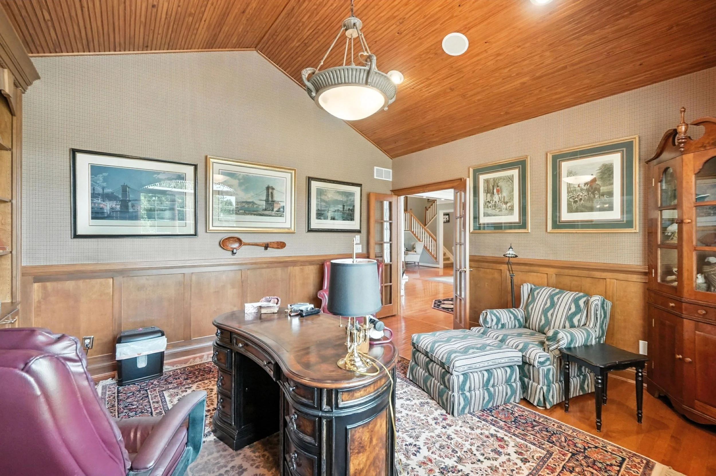 Living room with wood-paneled ceiling, framed artwork, wooden furniture, a patterned rug, a striped sofa, and a desk with a black lamp, with a view into a hallway with stairs.