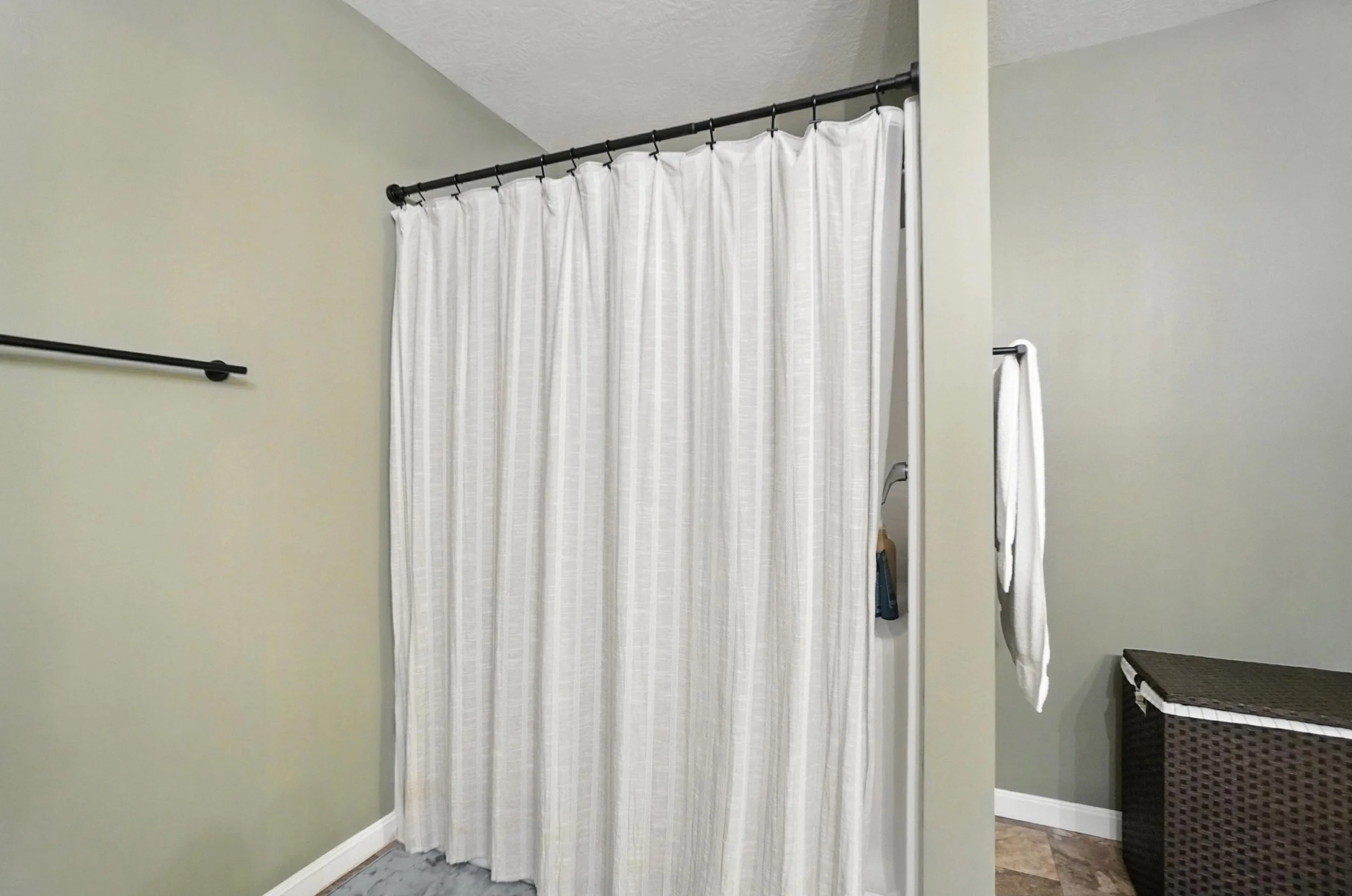 Indoor shower area with white curtains, a brown wicker storage basket, and a white towel hanging on a black hook.