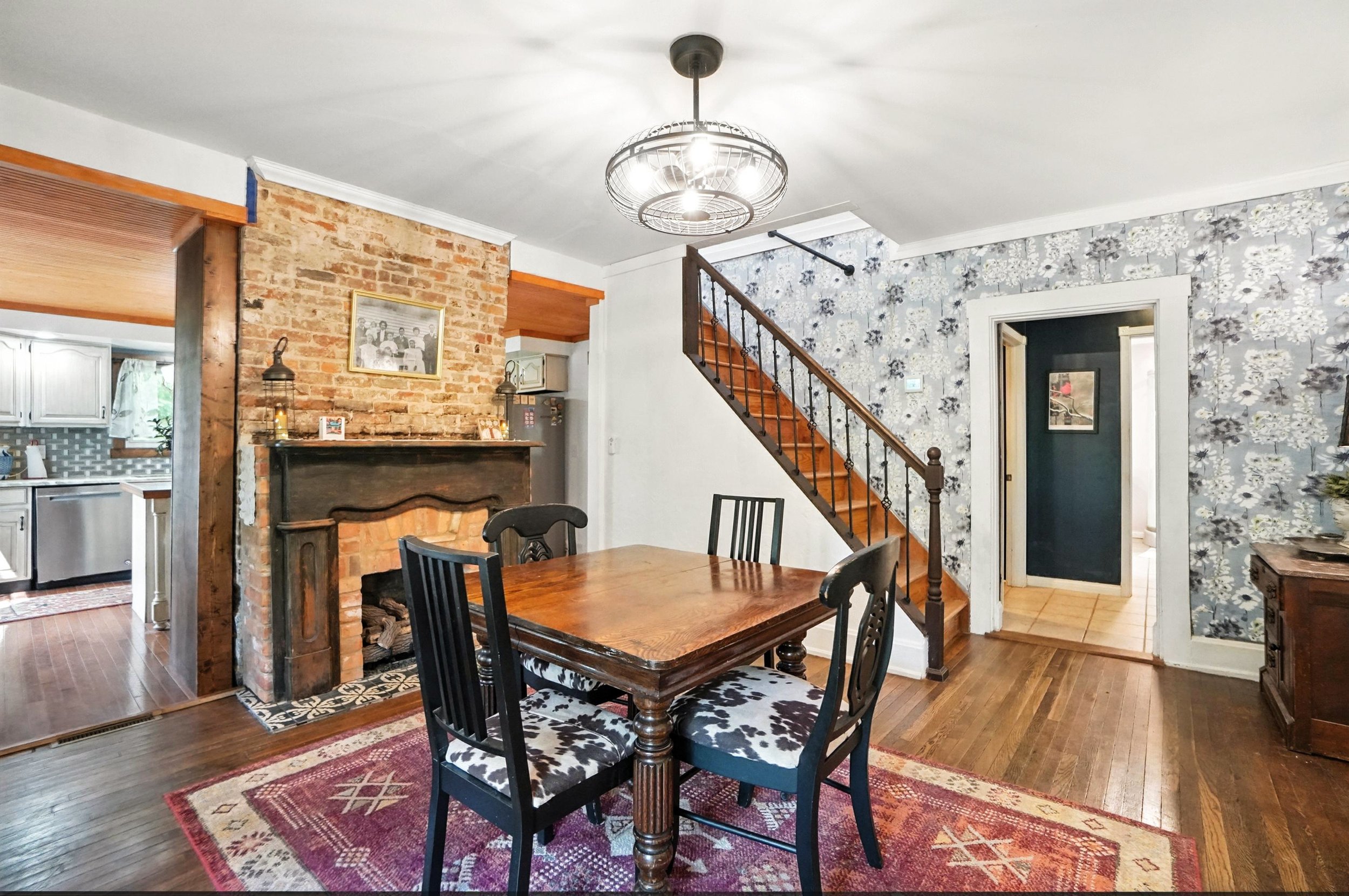 A cozy dining room with a wooden table and four chairs. A brick fireplace with a wooden mantle is on the left. A staircase with wooden steps and black metal railing ascends in the background. The room has hardwood floors, a patterned area rug, floral