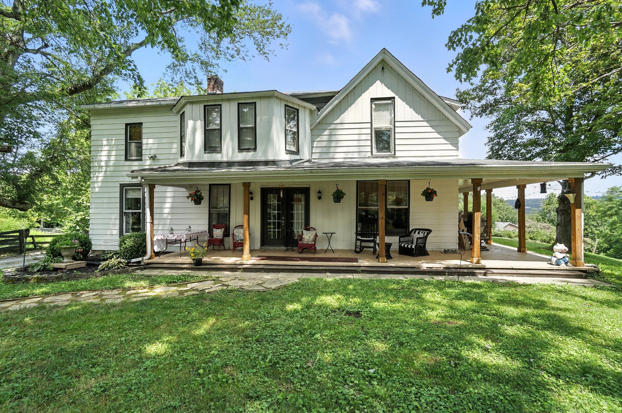 White two-story house with a large covered porch, wooden posts, outdoor furniture, and hanging flower baskets, surrounded by green grass and trees.