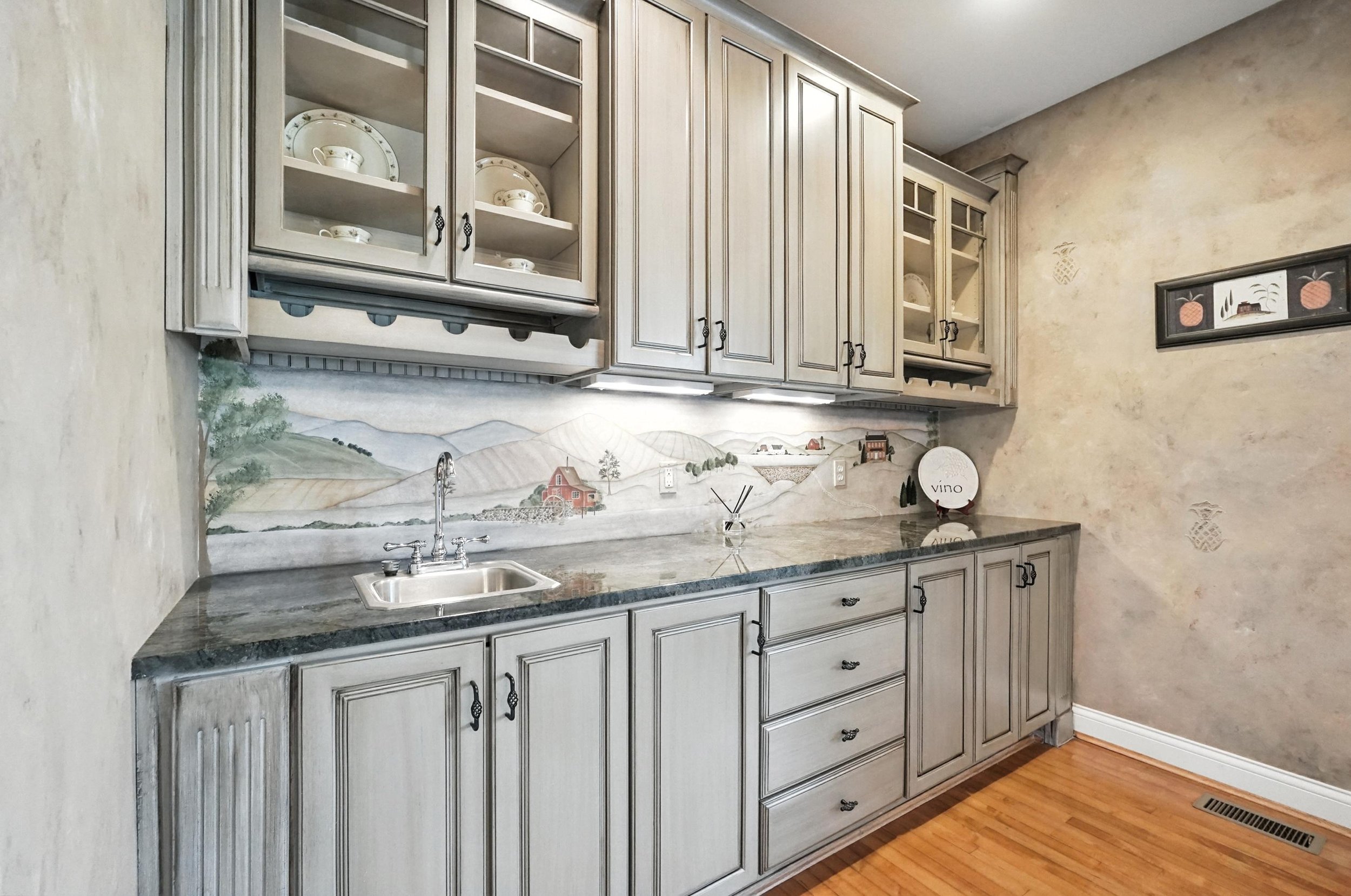 Kitchen with gray cabinets, a small sink, a granite countertop, decorative dishes, and a landscape mural backsplash.