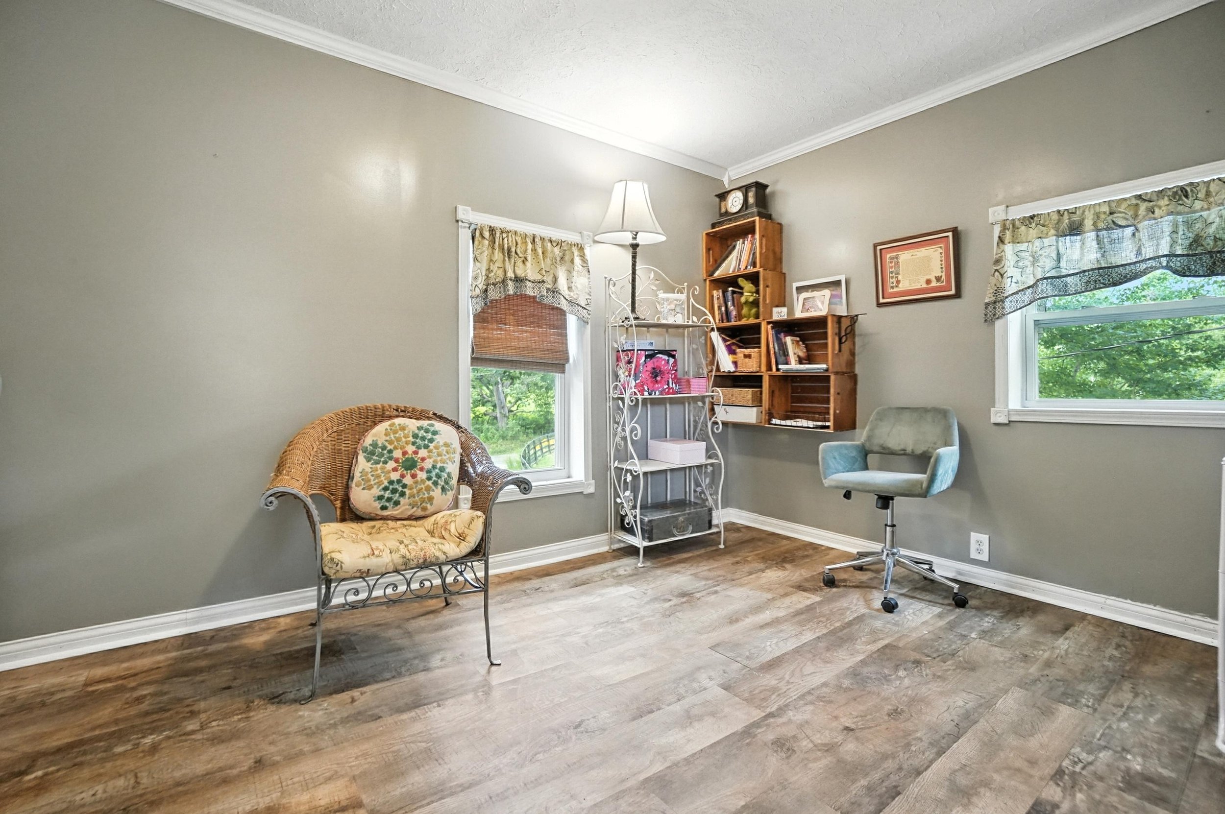 A room with two windows, green walls, wooden flooring, and a mix of vintage and modern furniture, including a wicker armchair with a floral cushion, a green swivel chair, an ornate white metal floor lamp, and a wooden bookshelf filled with books and