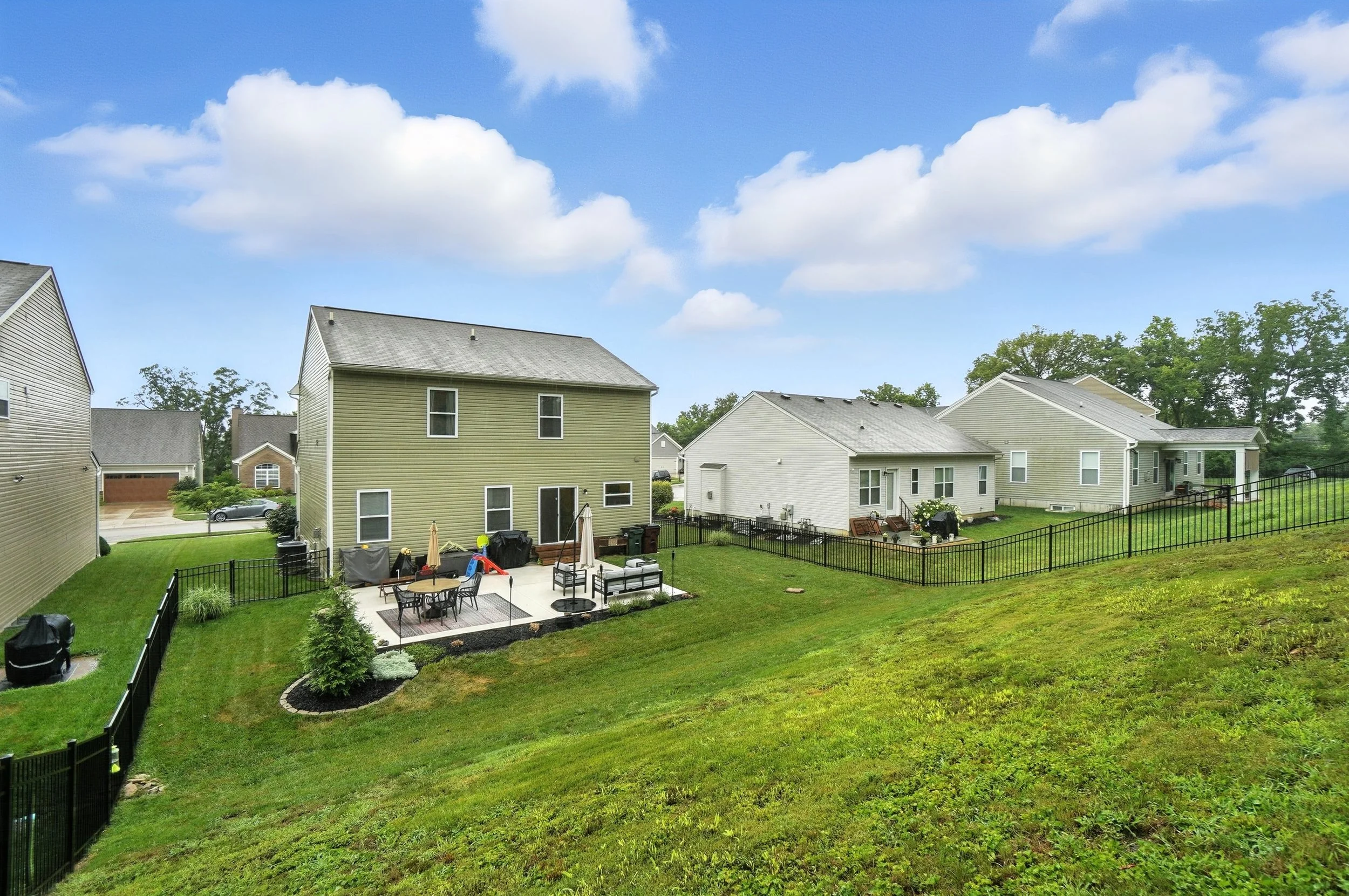 Residential backyard with patio furniture, umbrellas, grill, and lush green lawn surrounded by houses and a black chain-link fence under a partly cloudy sky.