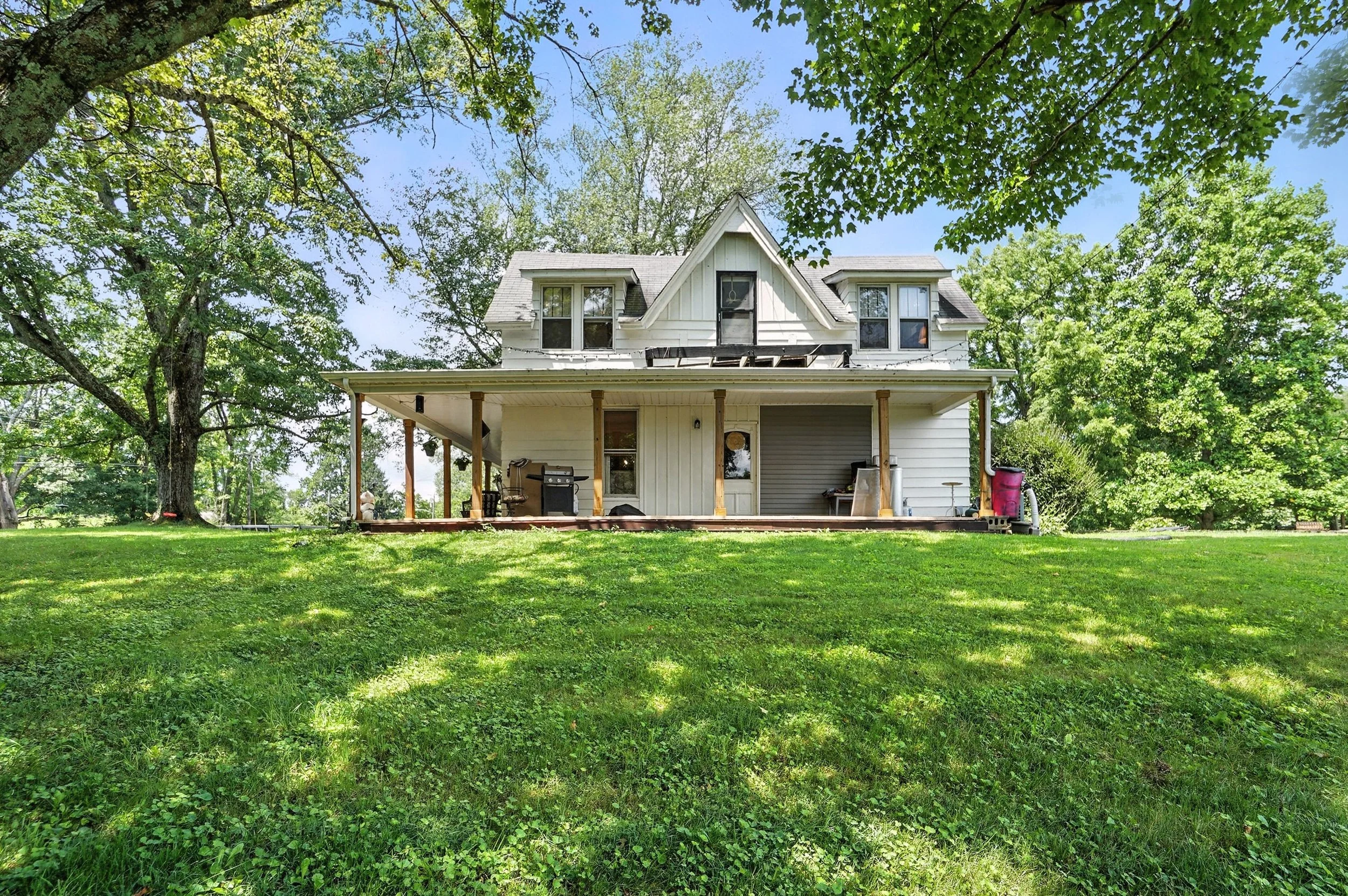 A two-story white house with a large covered porch on a grassy yard surrounded by trees.