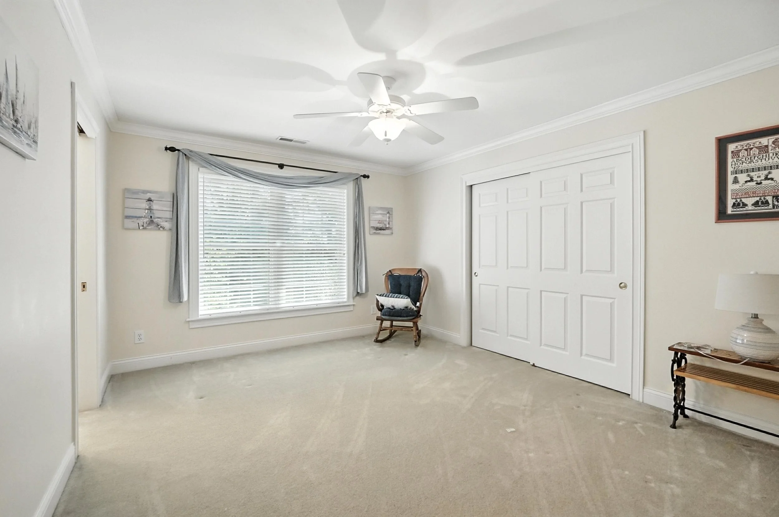 Empty bedroom with a large window, a ceiling fan, a wooden chair with cushions, a small side table with a lamp, and framed pictures on the walls.