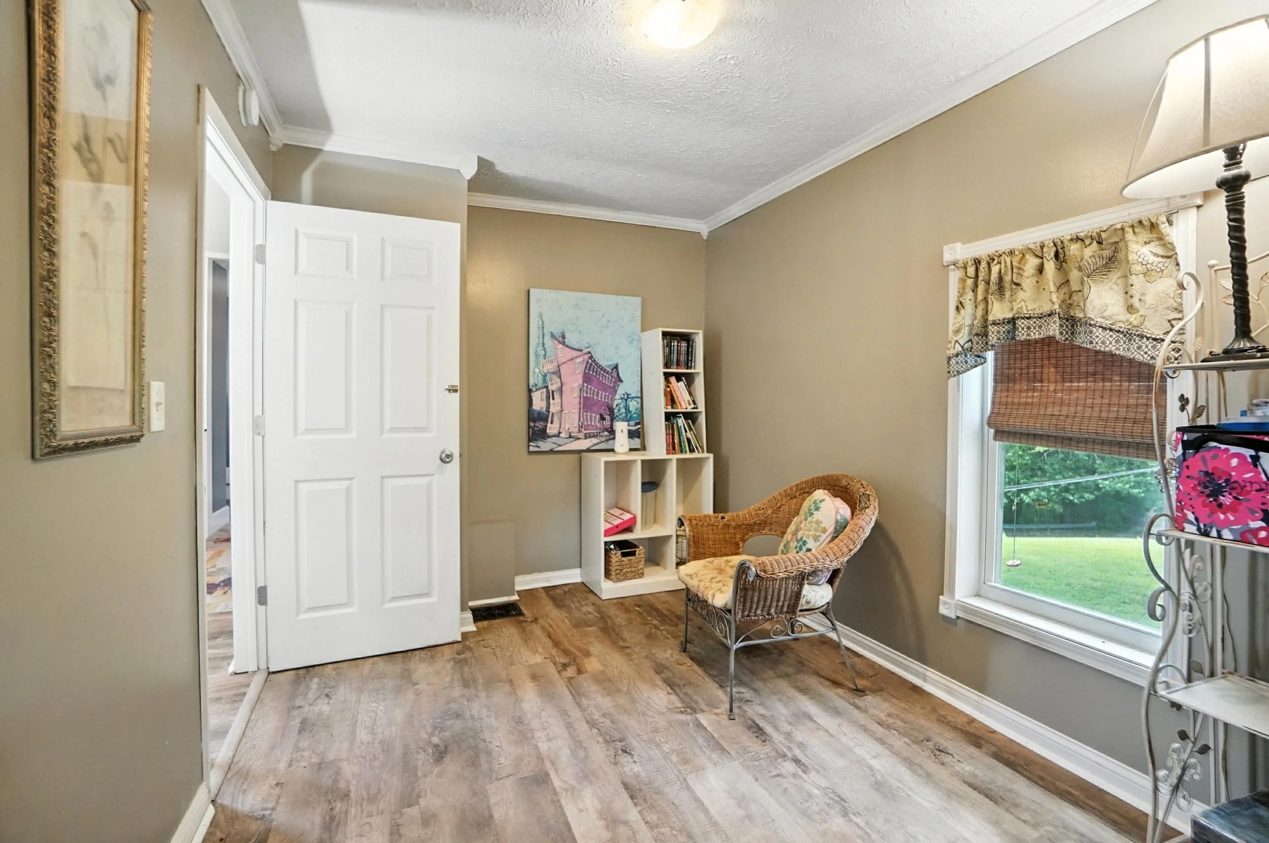 A cozy living room corner with beige walls, a large window with floral and bamboo blinds, a wicker chair with floral cushions, a small white bookshelf with books, a colorful painting on the wall, and a lamp with a beige shade.