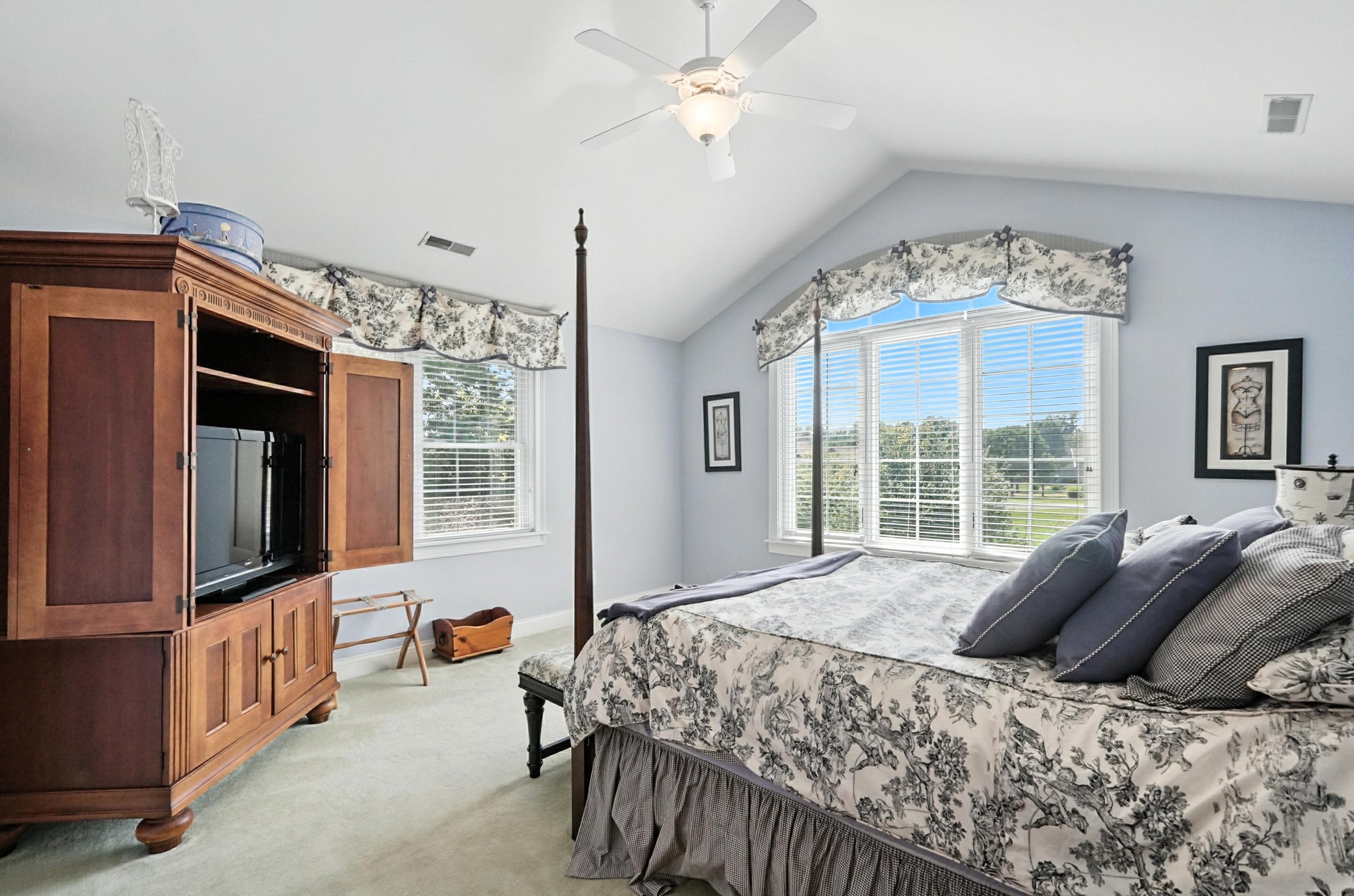 Bright bedroom with a large bed with patterned bedspread and multiple pillows, a wooden entertainment center with a TV, two windows with floral valances, and a ceiling fan.