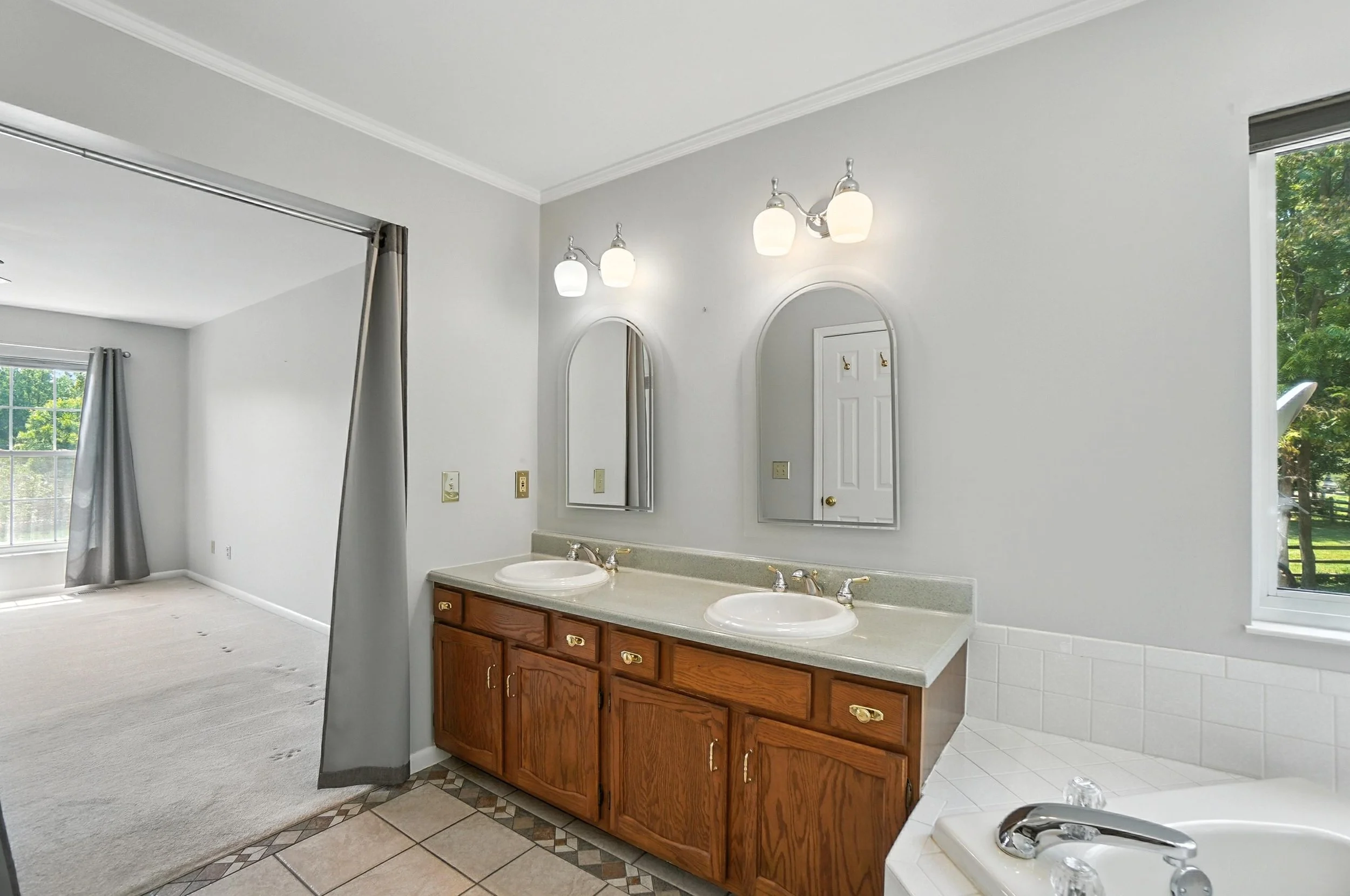 Bathroom with double sink vanity, two mirrors, wall-mounted light fixtures, and a bathtub with a window showing green trees outside.