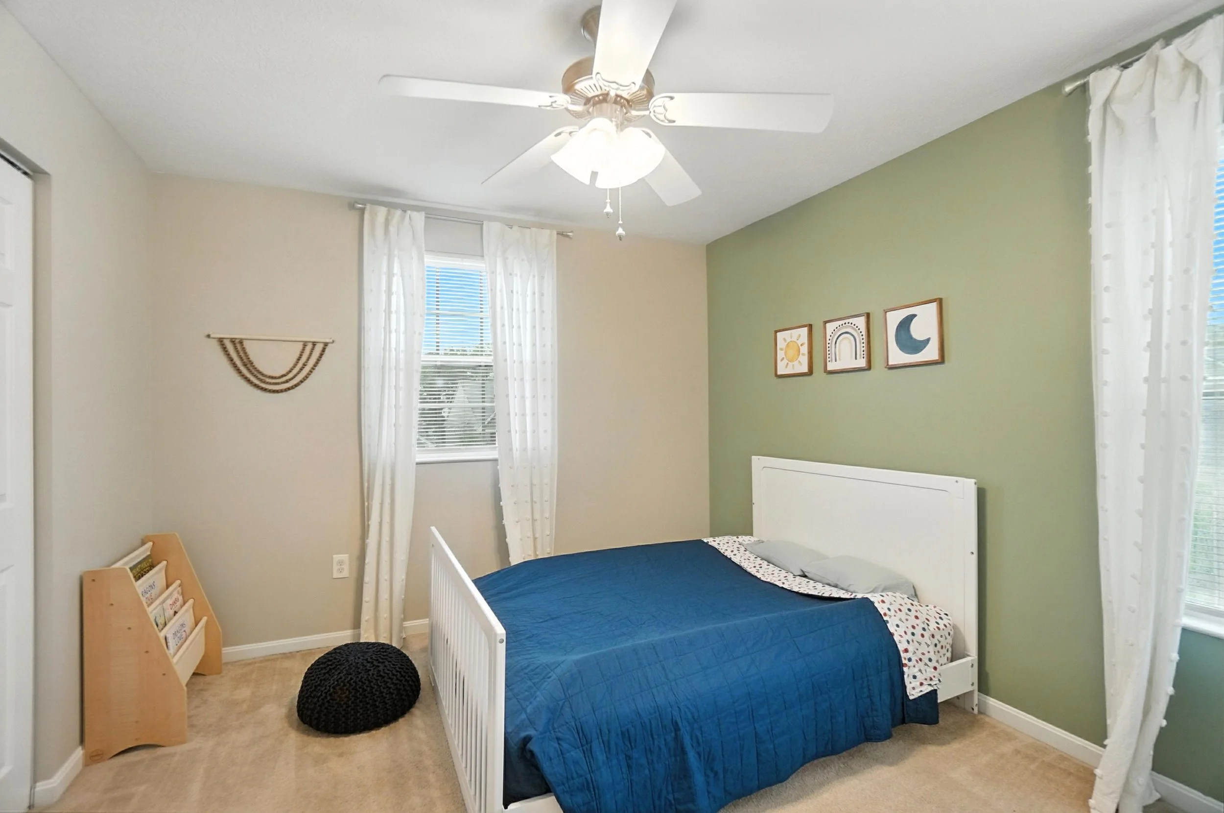 A bedroom with a white bed, navy blue quilt, and gray pillows. Green accent wall with three framed pictures of a sun, rainbow, and moon. White curtains on windows, beige carpet, ceiling fan with light, and black knitted pouf on floor.