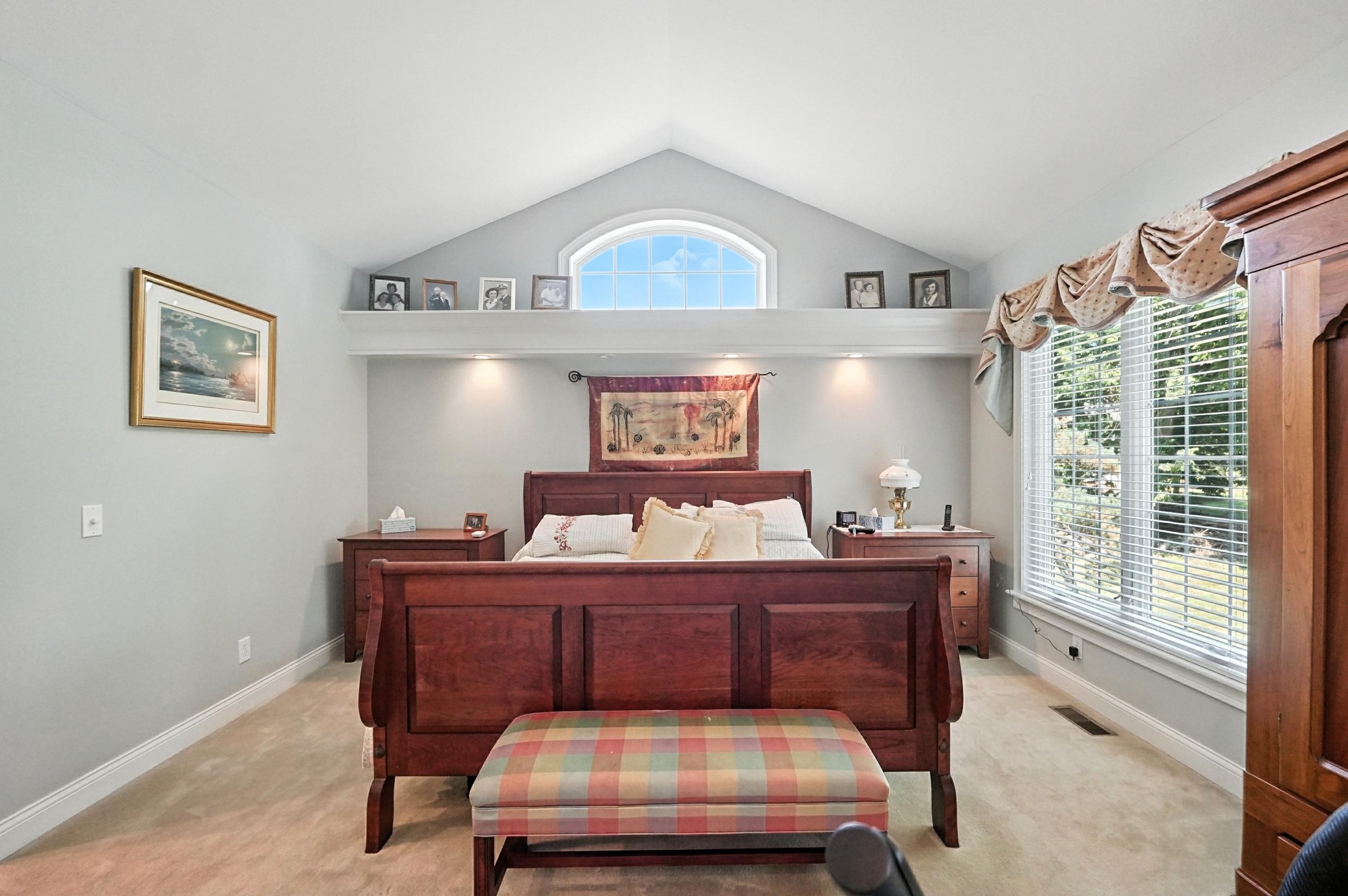 Bedroom with a wooden bed frame, matching nightstands, and a window with blinds and valance. Decor includes framed pictures and a tapestry above the bed, with a bench at the foot of the bed.