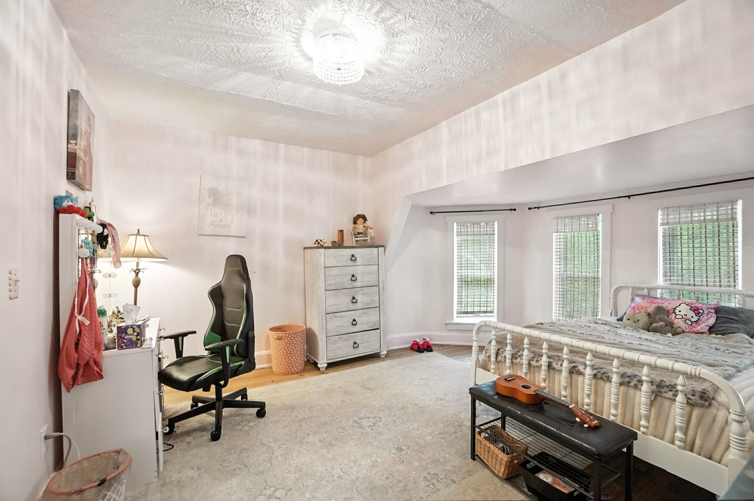 A bedroom with a bed near three windows, a white dresser, a black gaming chair, and a desk with items, featuring light-colored walls and a ceiling light fixture.