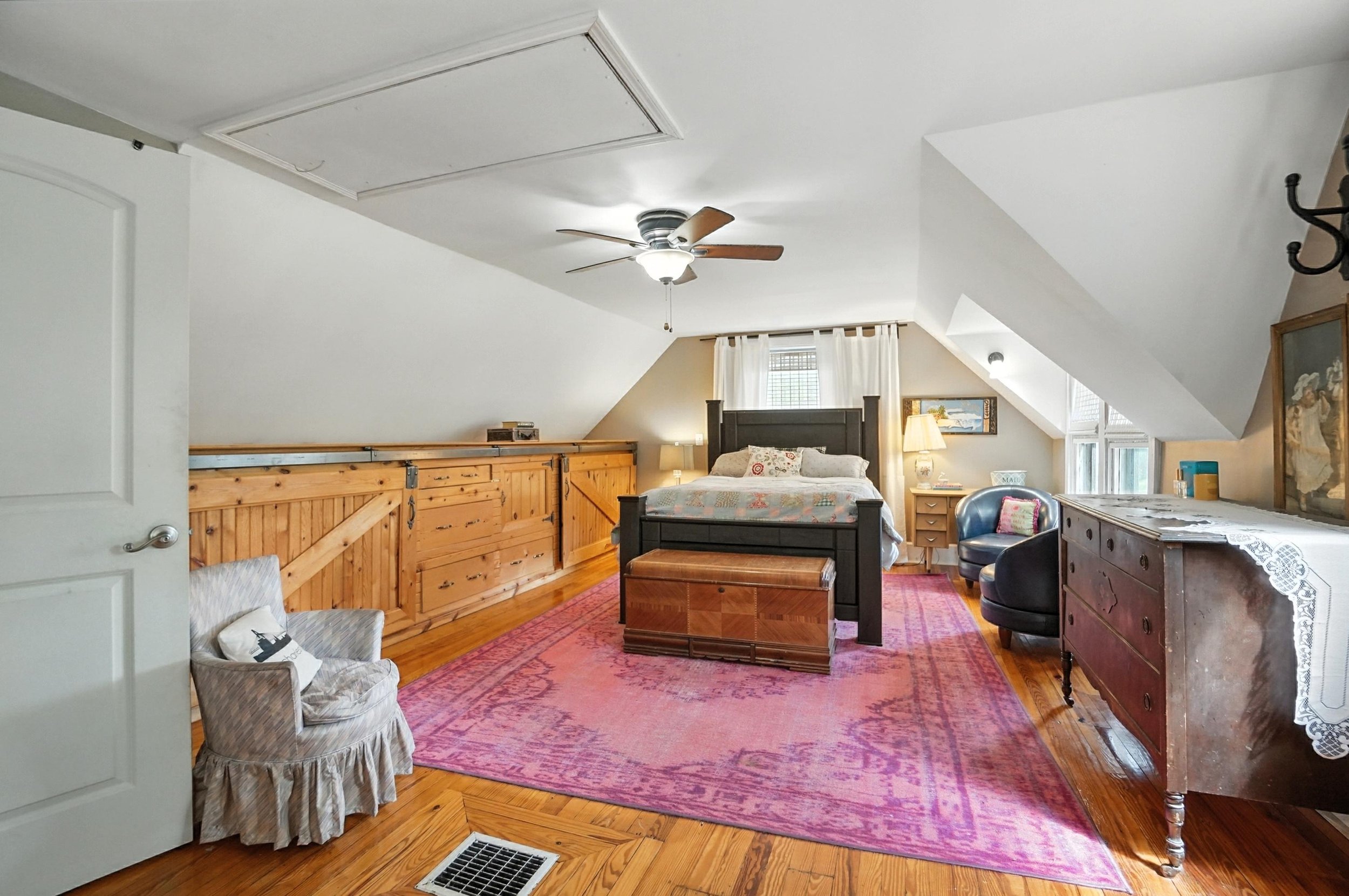 A cozy attic bedroom with a black bed, wooden furniture, a pink rug, and various chairs and lamps.
